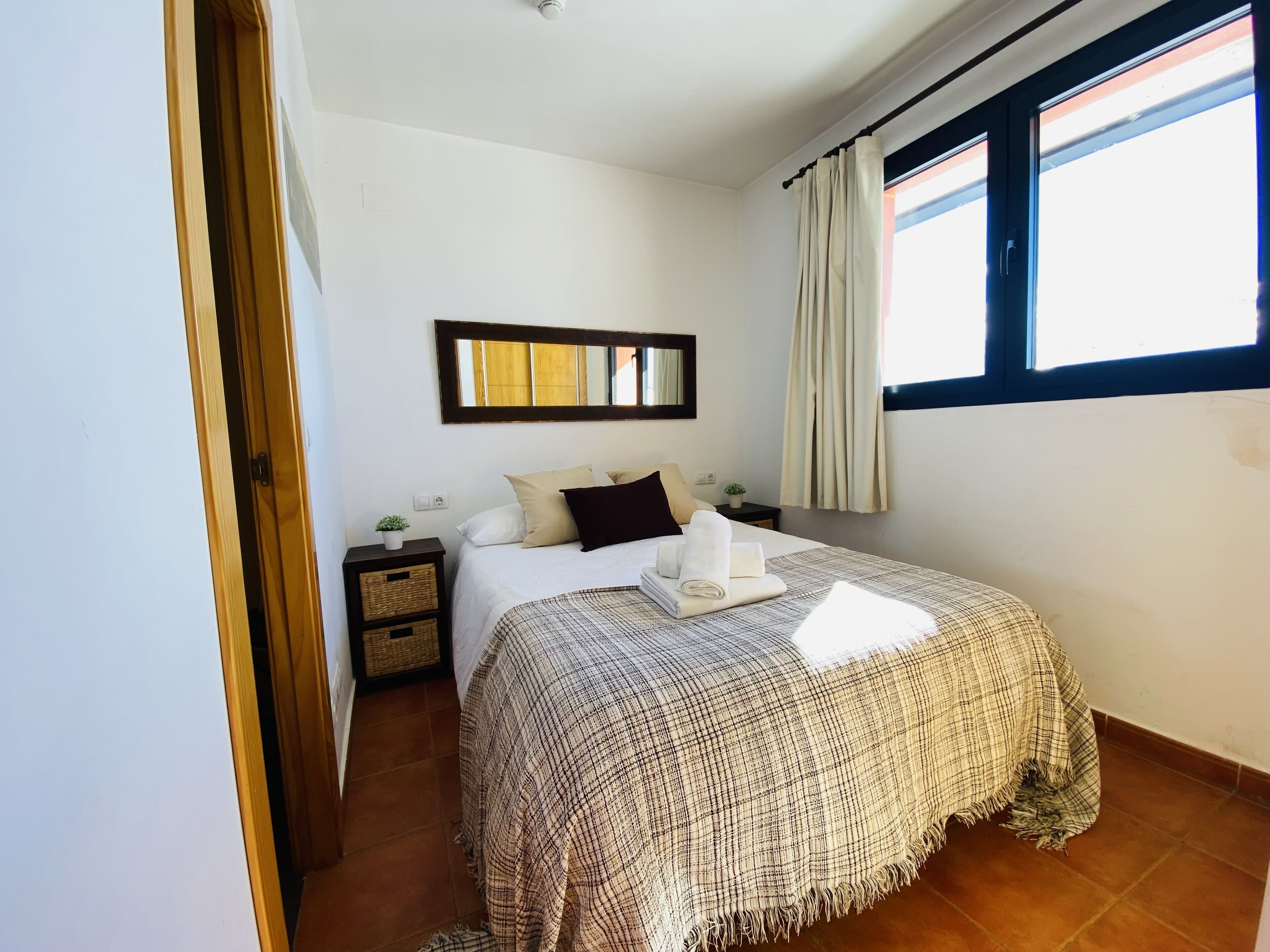 Un dormitorio con cama, mesita de noche, espejo, ventana con cortinas y puerta.