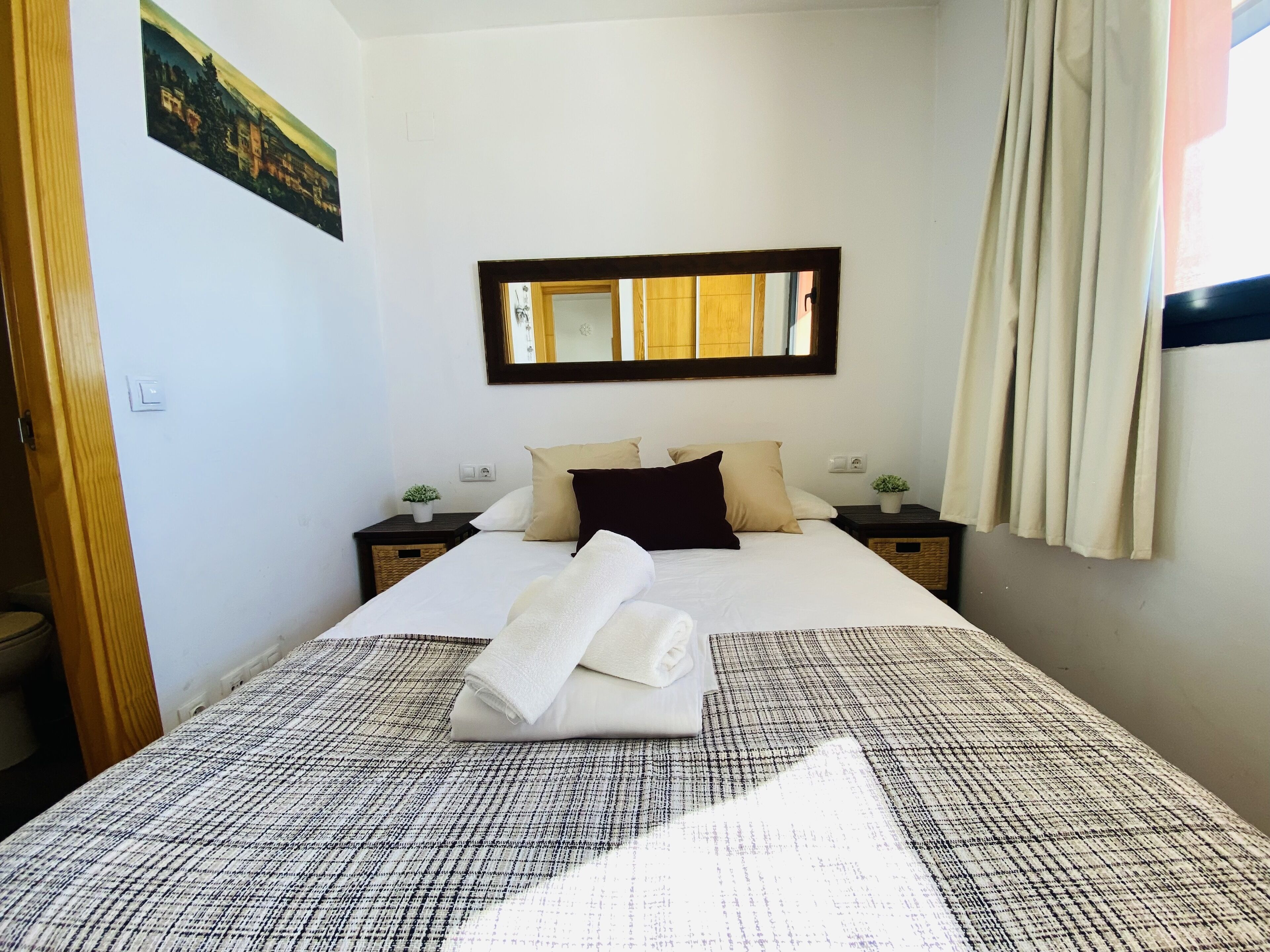 Una cama bien hecha con colcha a cuadros blancos y negros, dos mesitas de noche con pequeñas macetas y un espejo grande sobre la cama, además de una ventana con cortinas.