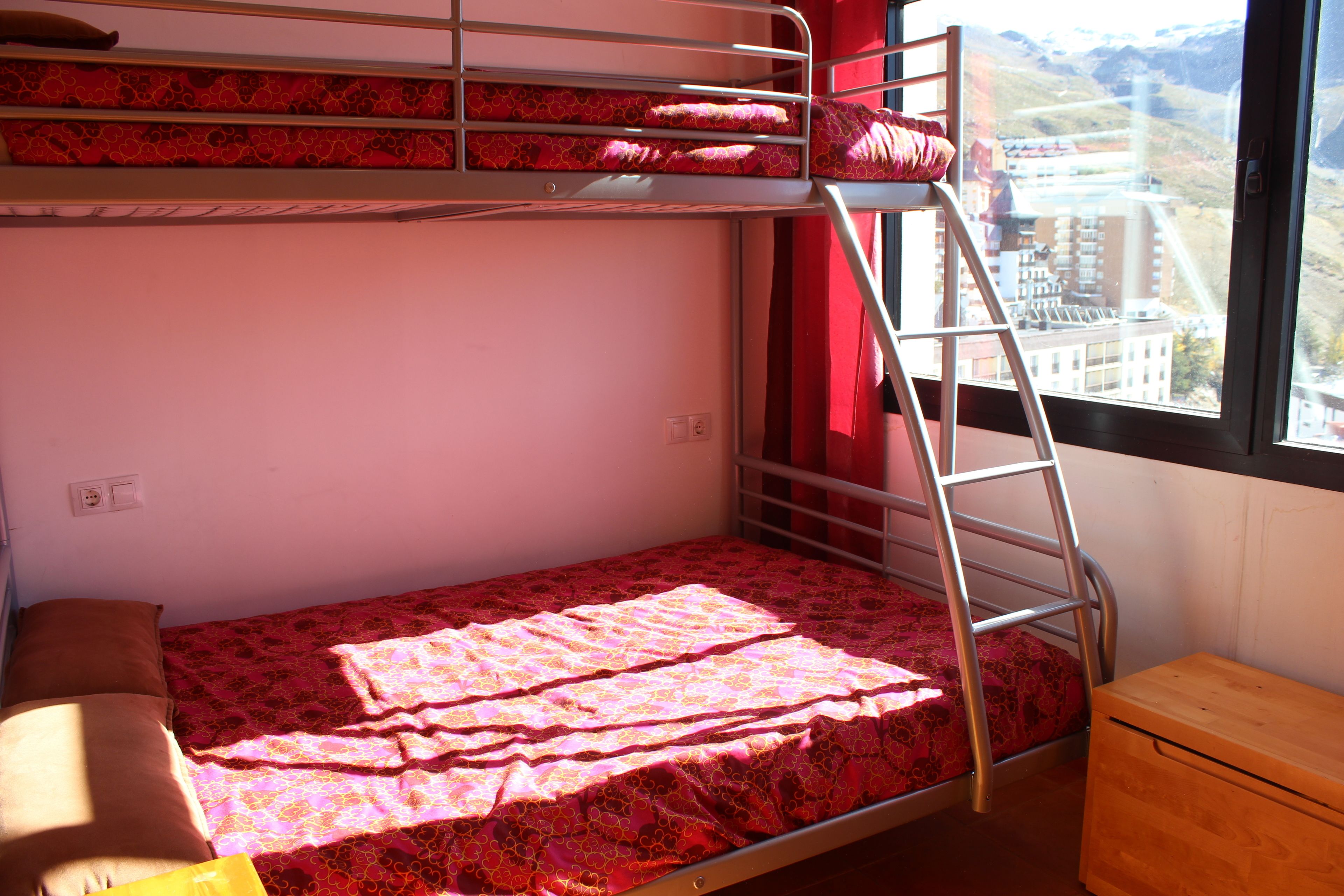A bunk bed with a ladder, a wooden nightstand, and a window with a view of buildings.