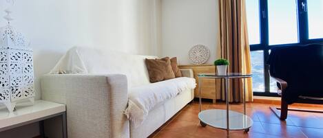 A modern living room with a white sofa, a small round table, and a view of the city through large windows.