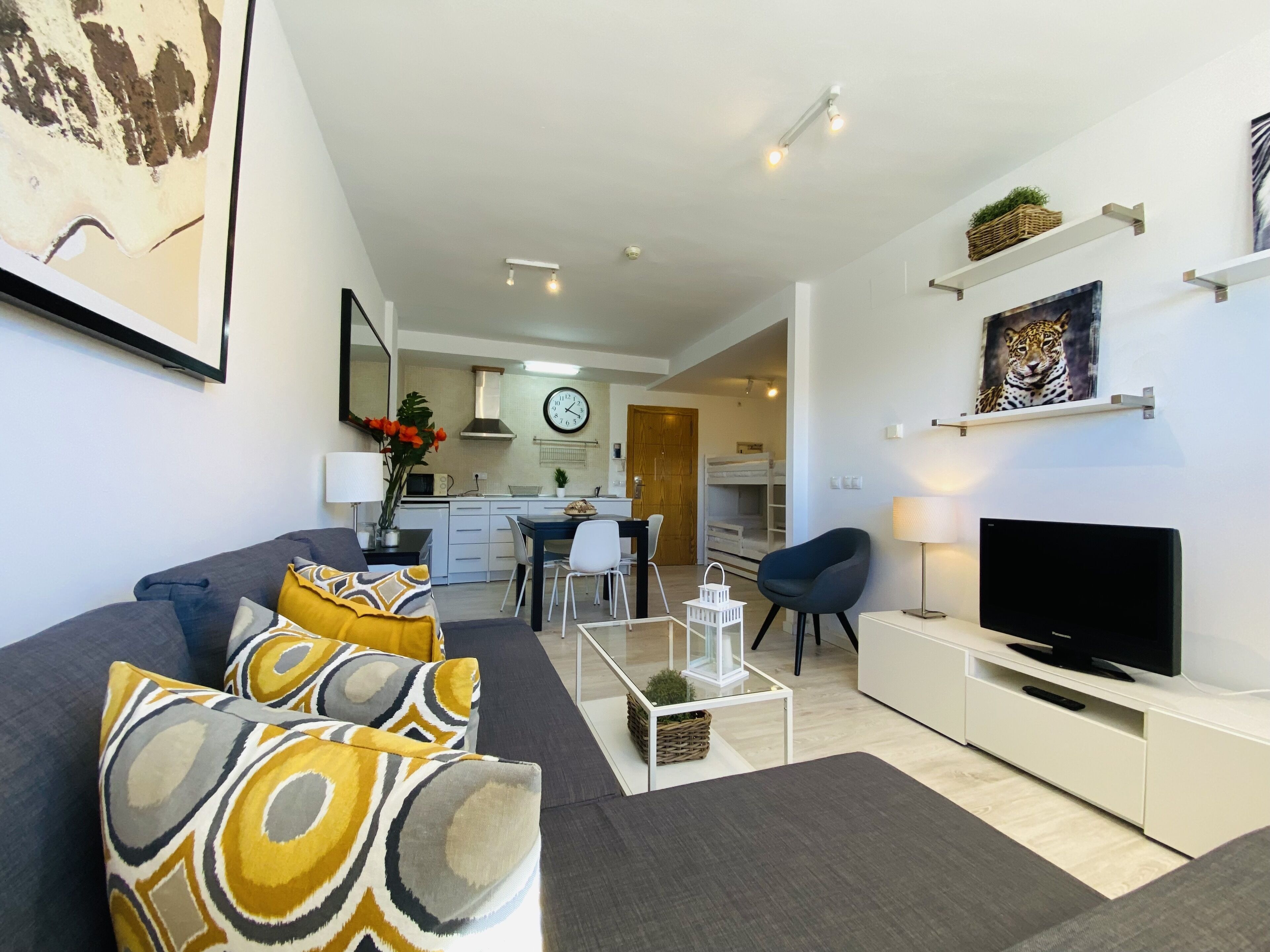 Una sala moderna con sofá, televisor y comedor, con una cocina al fondo.