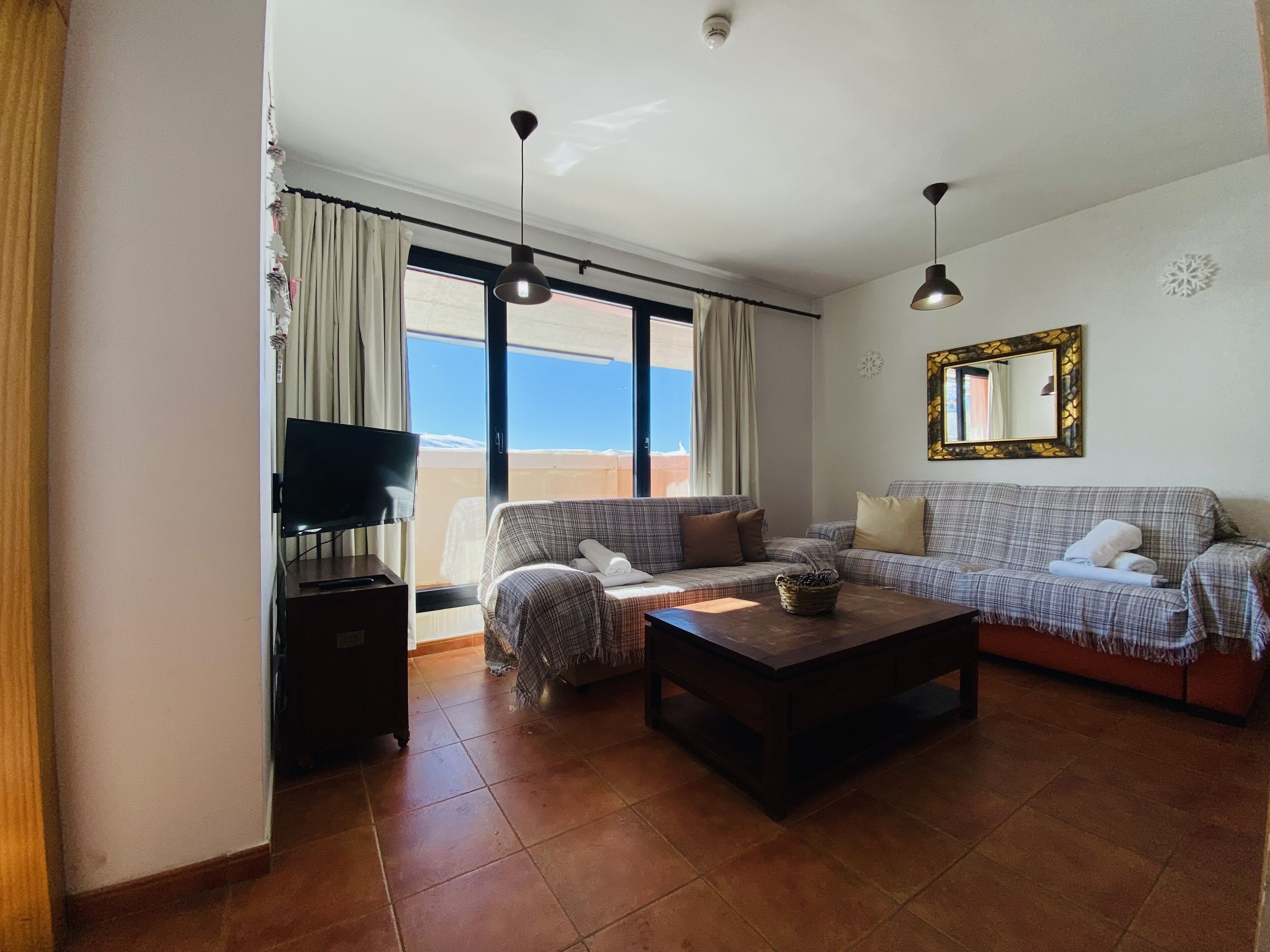 Una sala de estar con sofá, mesa de centro y televisor. Se ve una ventana con cortinas y vistas al exterior.