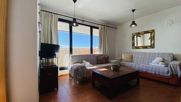 Una sala de estar con sofá, mesa de centro y televisor. Se ve una ventana con cortinas y vistas al exterior.