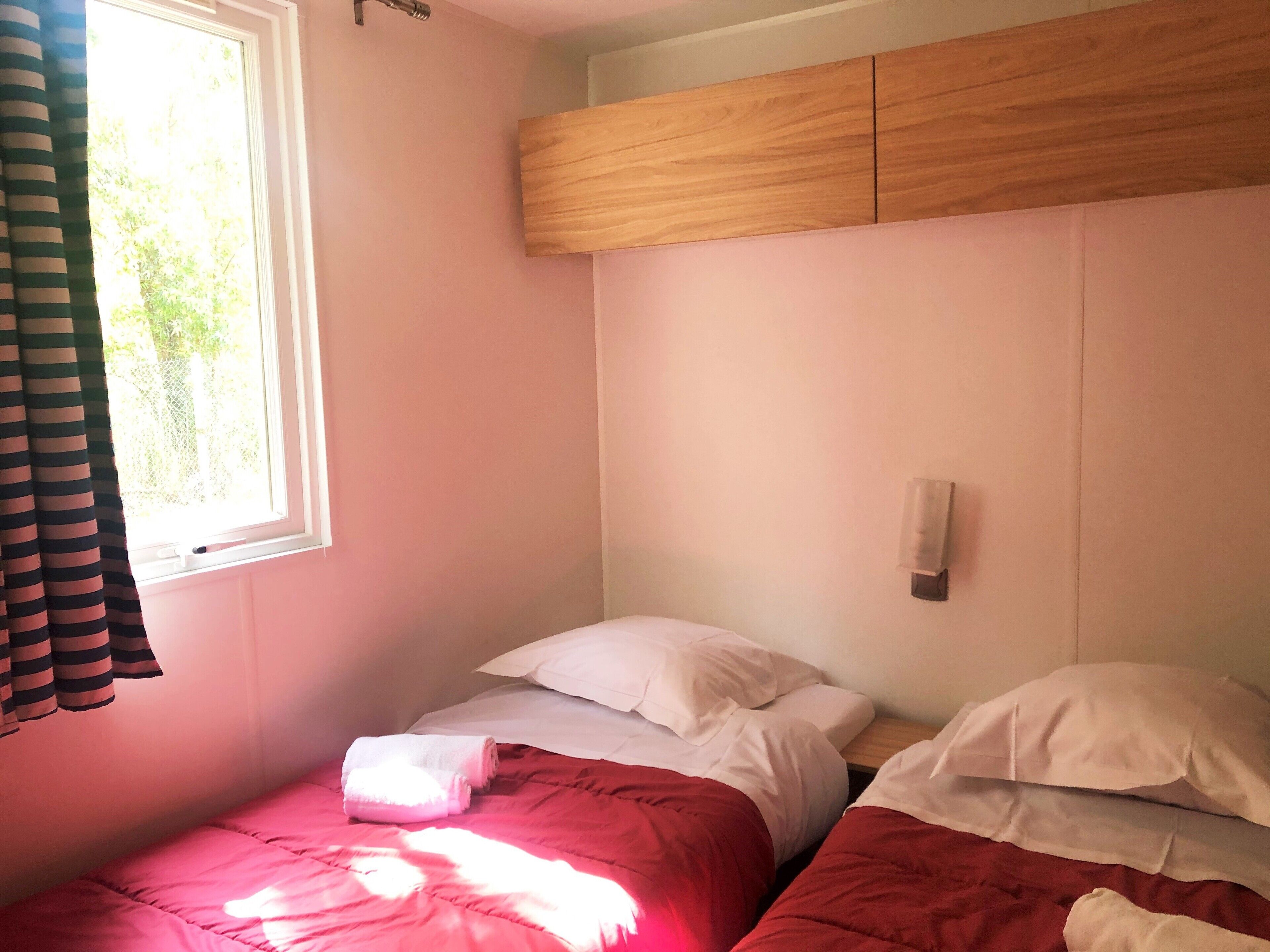 Una habitación con dos camas, cada una con almohadas blancas y ropa de cama roja. Se ve una ventana con cortina a la izquierda, y un panel de madera sobre las camas.