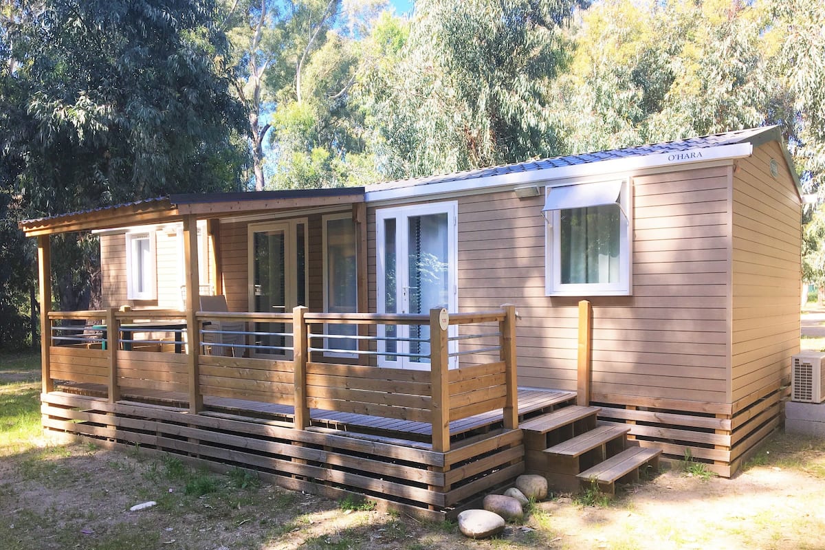 A small wooden cabin with a deck and a covered porch, surrounded by trees.