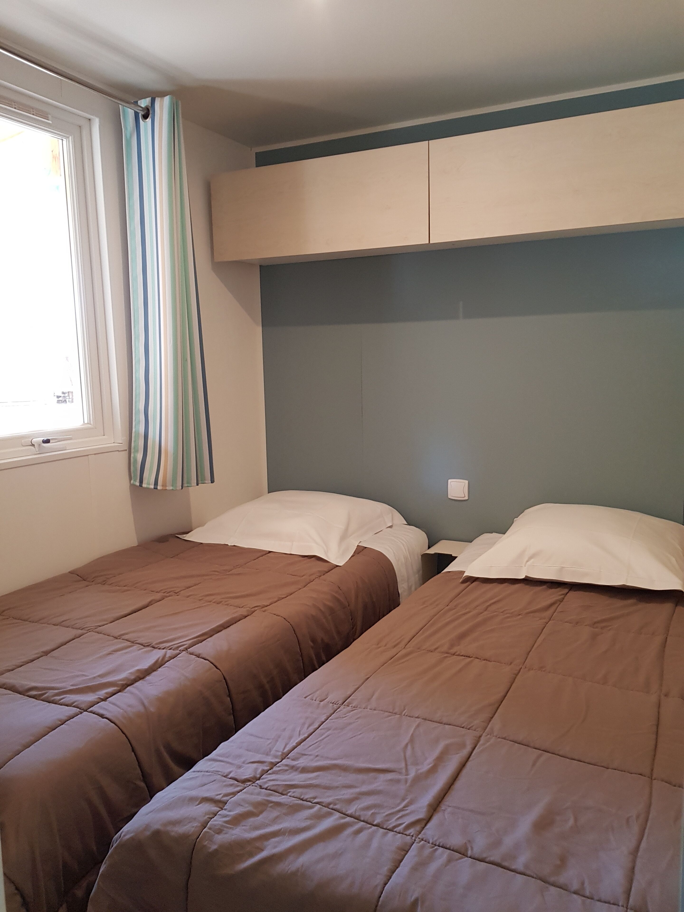 A room with two single beds, each with a brown bedspread and white pillows. There is a window with a striped curtain, and the walls are painted in a light blue and white color scheme.