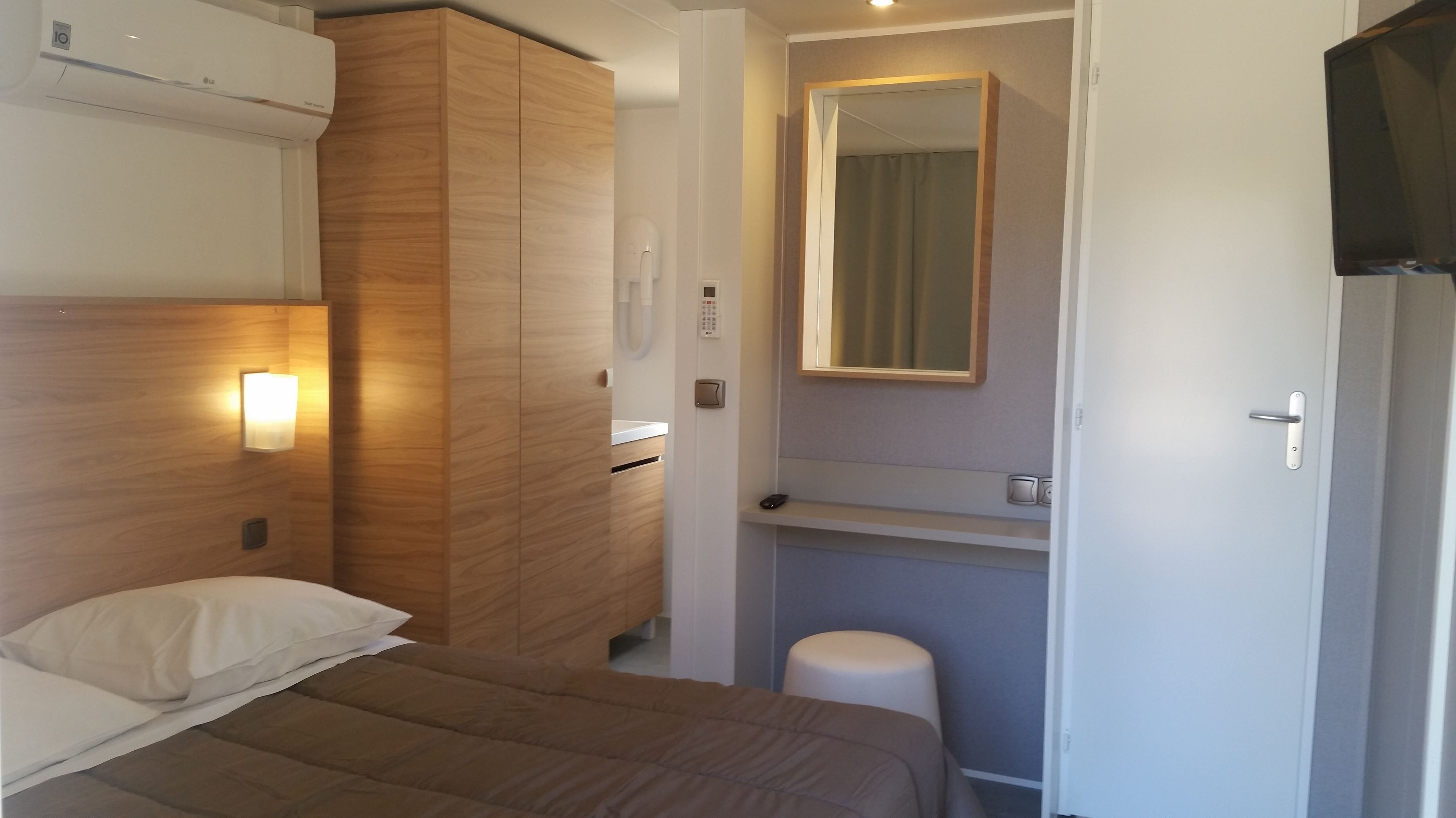 Una habitación de hotel con cama, un armario de madera, un taburete pequeño, un espejo y un televisor.