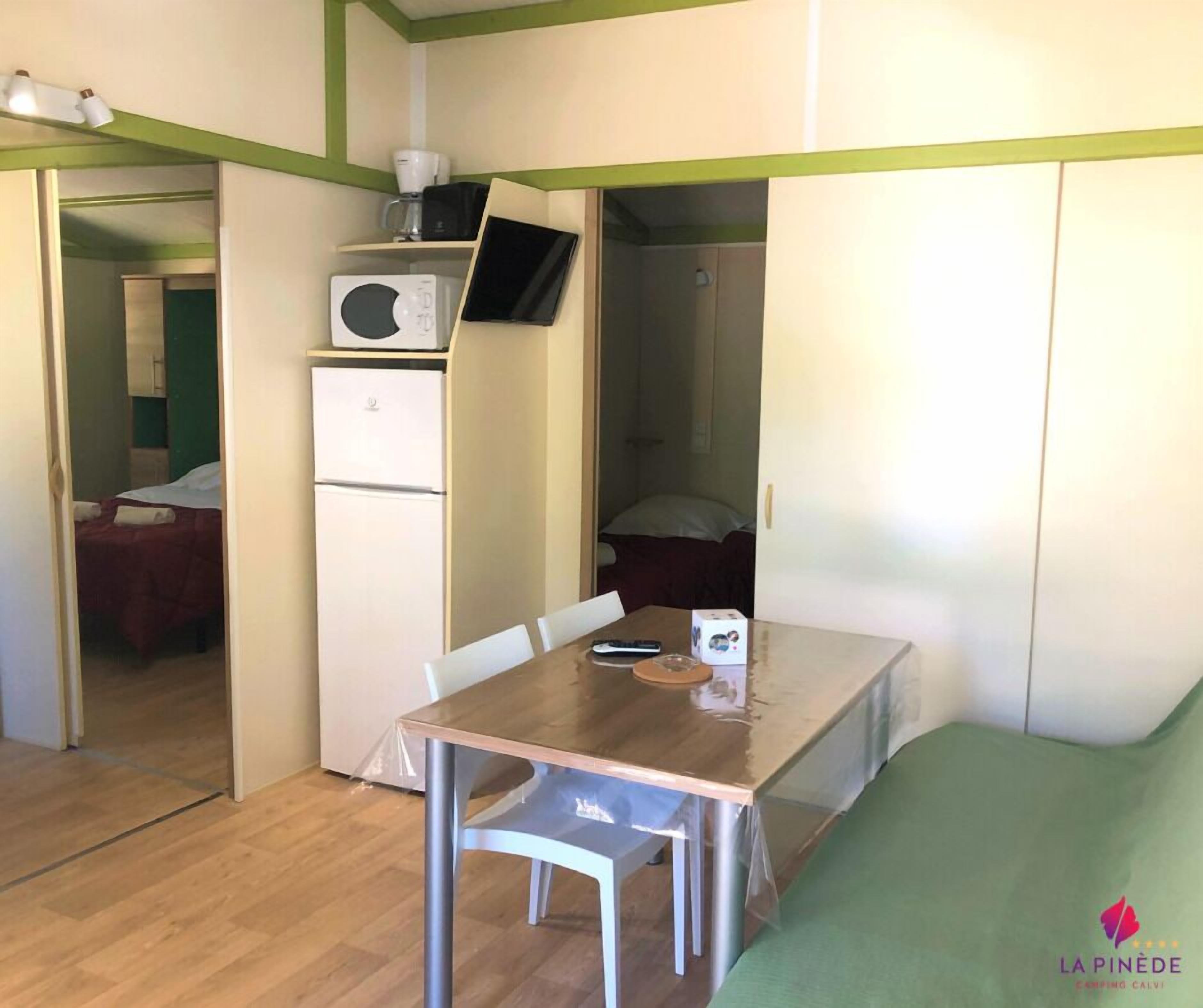 A compact room with a wooden table, a white refrigerator, a microwave, and a bed with a green bedspread.