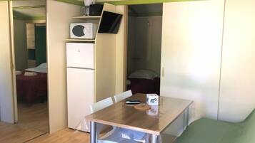 A compact room with a wooden table, a white refrigerator, a microwave, and a bed with a green bedspread.