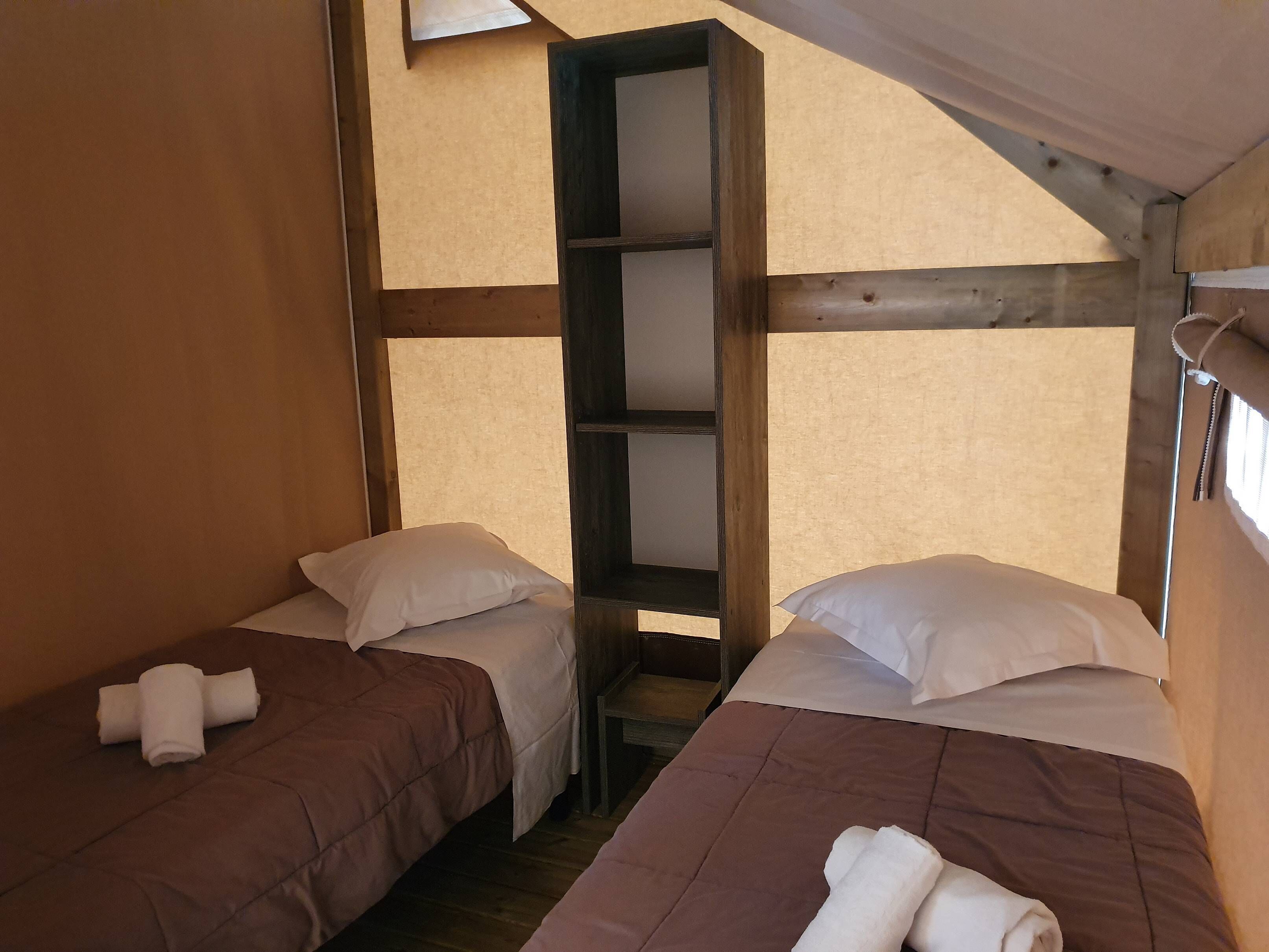 Une chambre avec deux lits, chacun étant équipé d’un oreiller et d’une serviette enroulée. Une étagère en bois est placée entre les deux lits.
