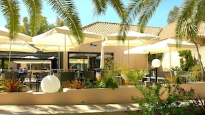 Un patio al aire libre con mesas y sillas bajo sombrillas, rodeado de palmeras y plantas en macetas.