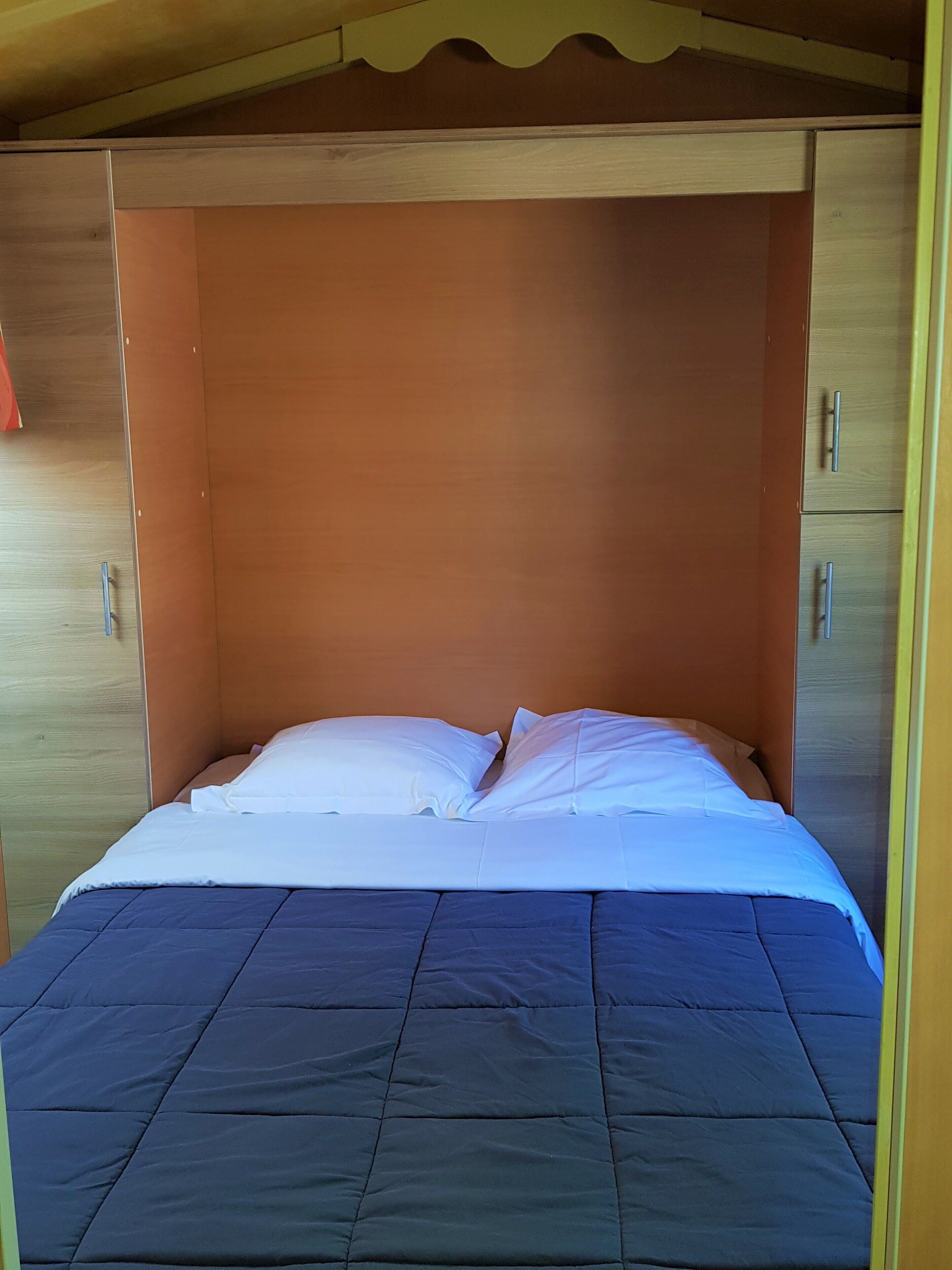Una cama con colcha azul y almohadas blancas en una habitación con paredes de madera y techo inclinado.
