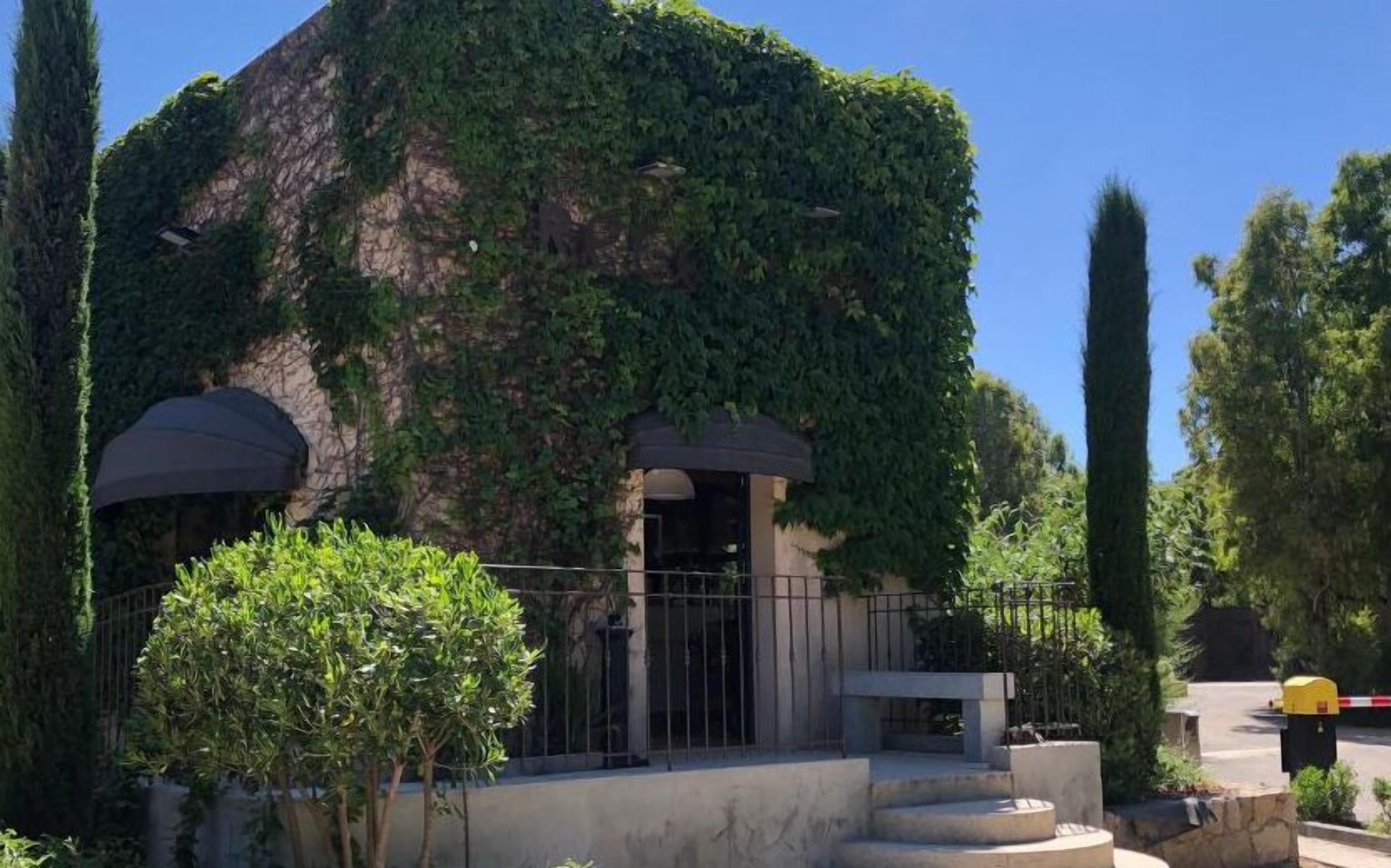 Un edificio con fachada de piedra parcialmente cubierta de hiedra, una pequeña entrada con una reja metálica y una escalera de hormigón que conduce a la entrada.