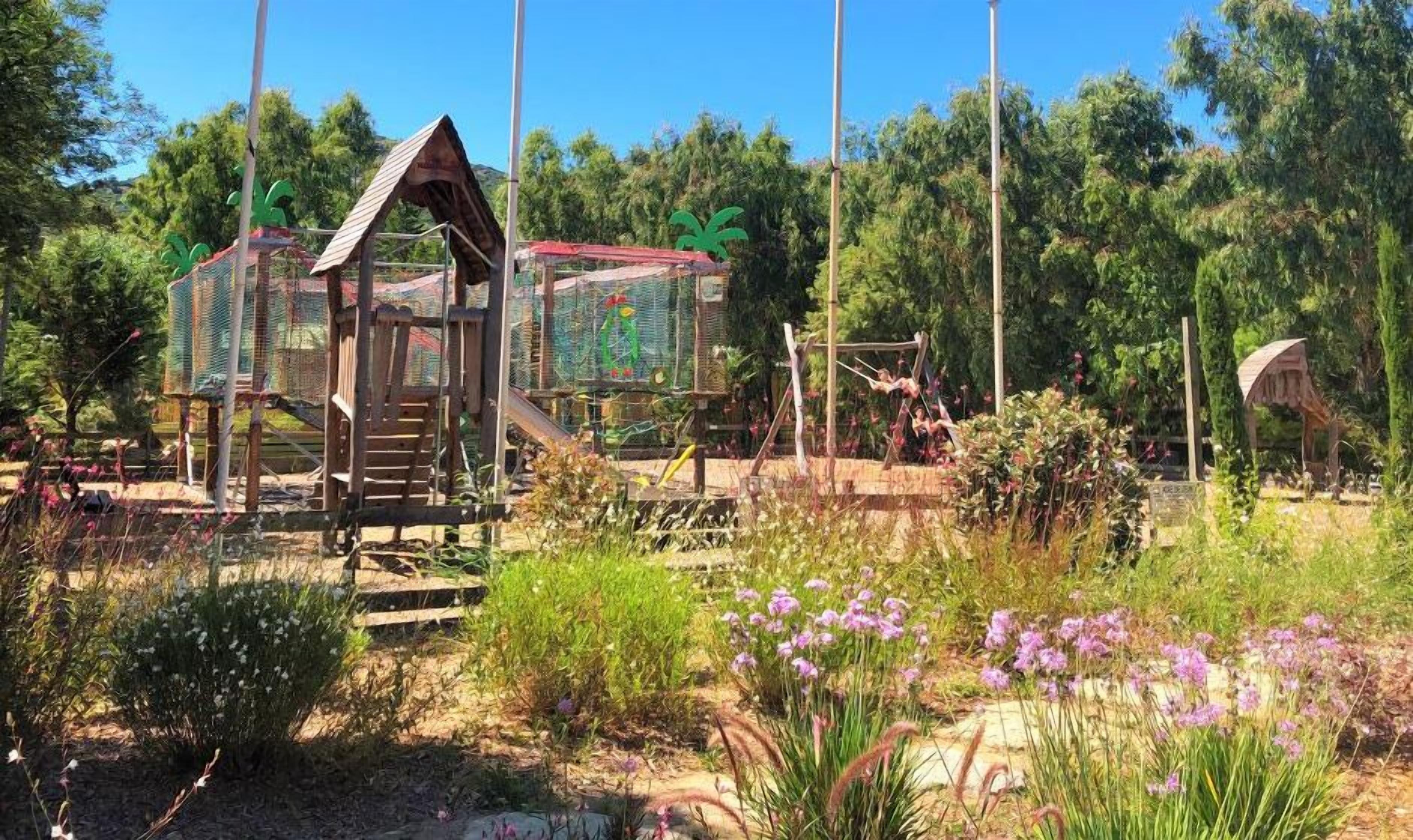 A wooden play structure with slides and climbing areas, surrounded by greenery and flowers.