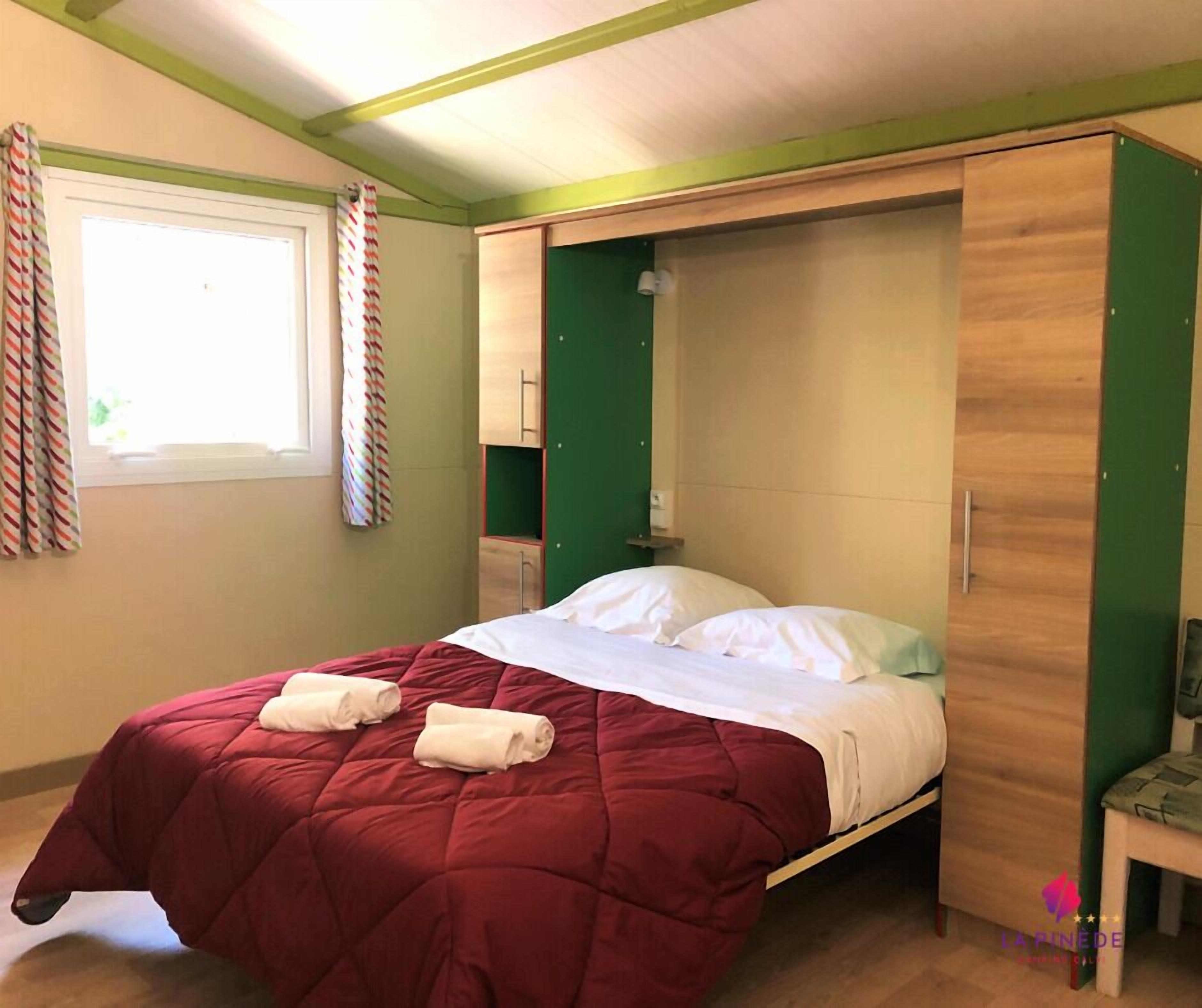 A single bed with a red quilt, a green wall, and a window with curtains.