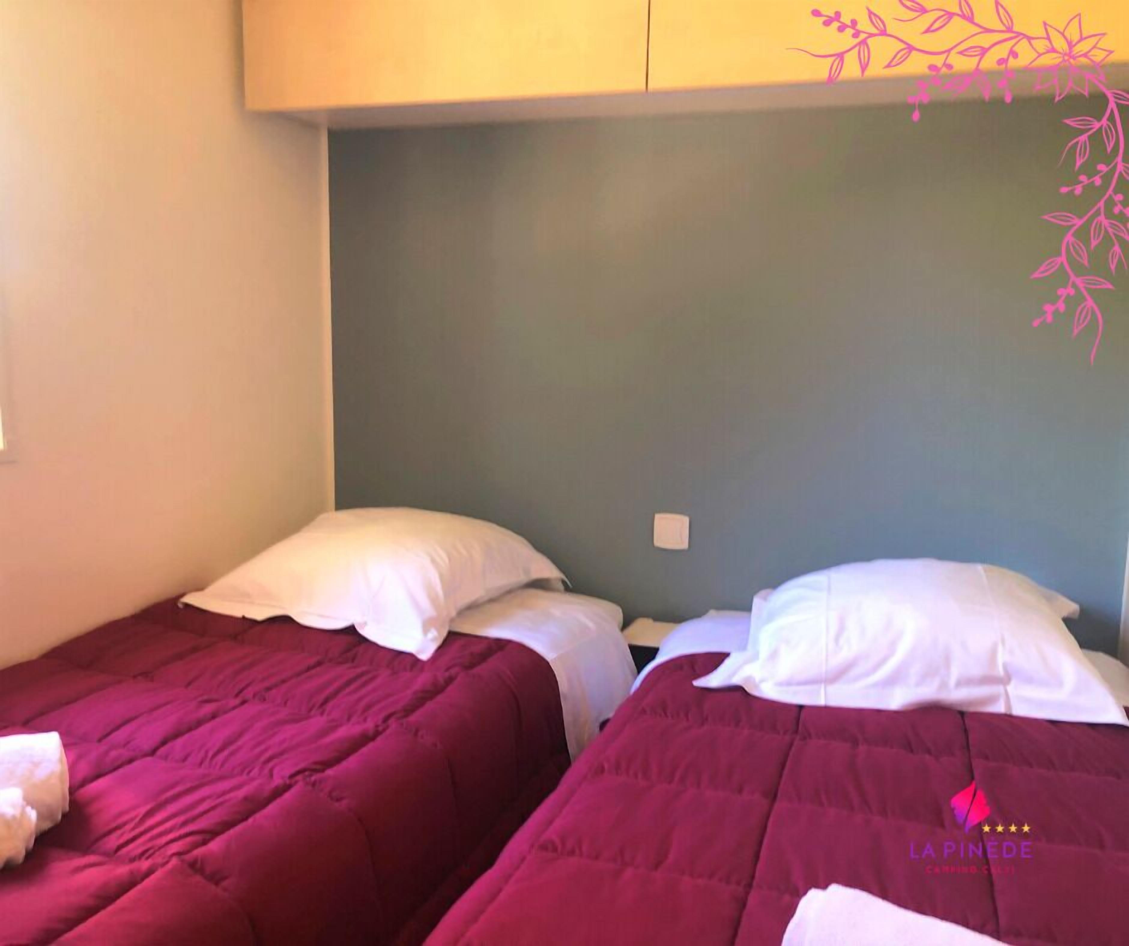Twee eenpersoonsbedden met witte kussens en een bordeauxrode sprei, een wandlamp en een decoratieve roze tak aan de muur.
