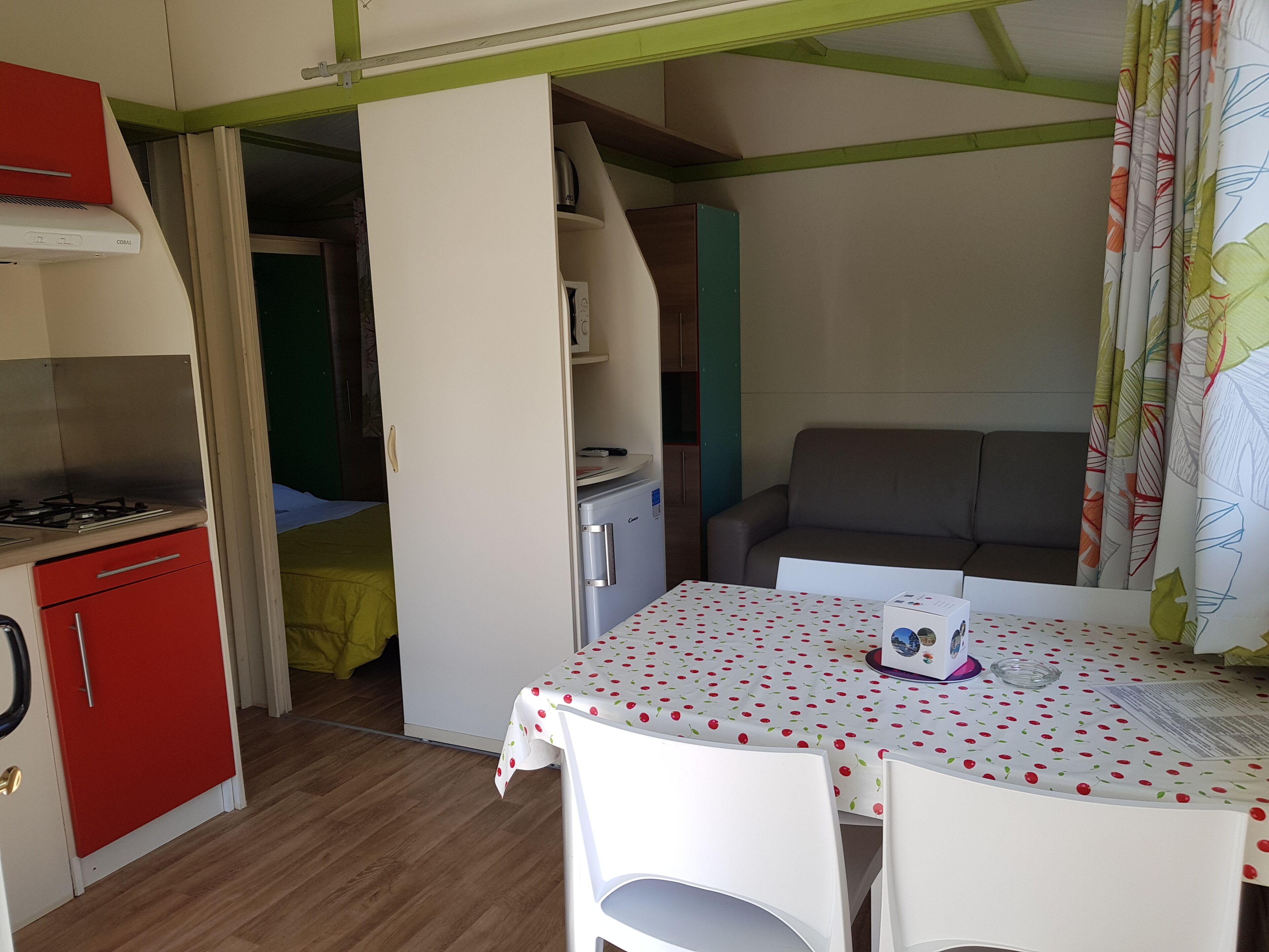 Une petite cuisine équipée avec un réfrigérateur rouge, une petite cuisinière et une table blanche recouverte d’une nappe à motifs de cerises.
