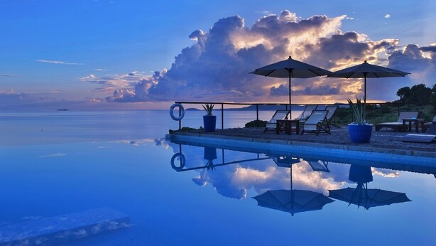 Una piscina infinita tranquila con sillones de descanso y sombrillas, que refleja el cielo y las nubes.
