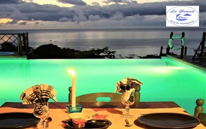 A dining table with a candle, plates, and cutlery set against a backdrop of a swimming pool and a view of the sea. - Le Grand Bleu (Nosy Be)
