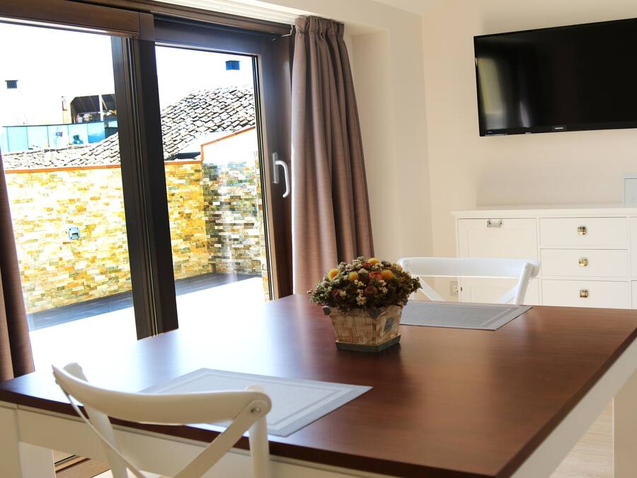 Een kamer met een houten tafel, witte stoelen, een flatscreen televisie aan de muur en uitzicht door een glazen deur op een buitenruimte met een betegeld dak.