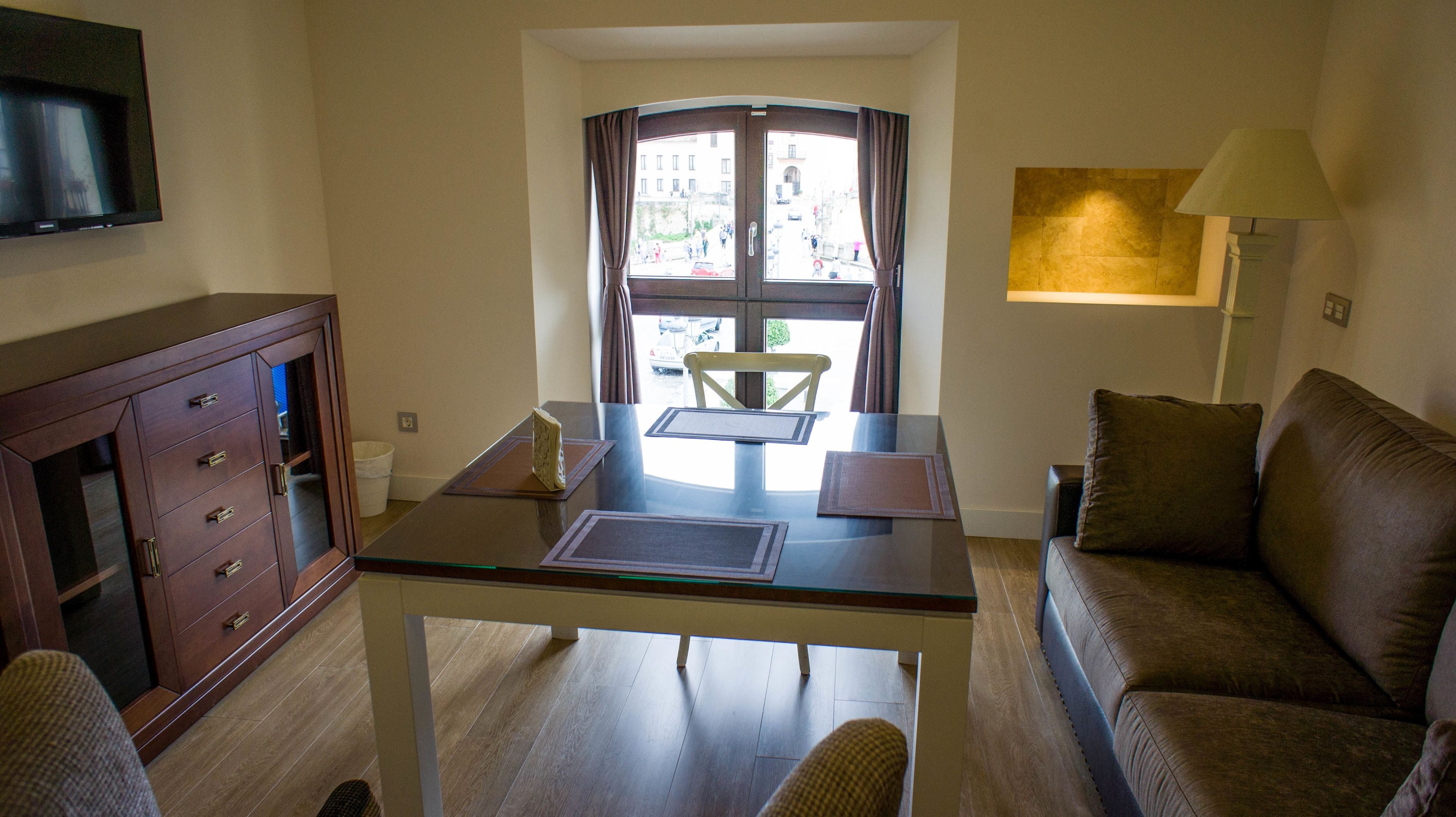 A living room with a glass table, a brown sofa, a TV mounted on the wall, and a window with curtains.
