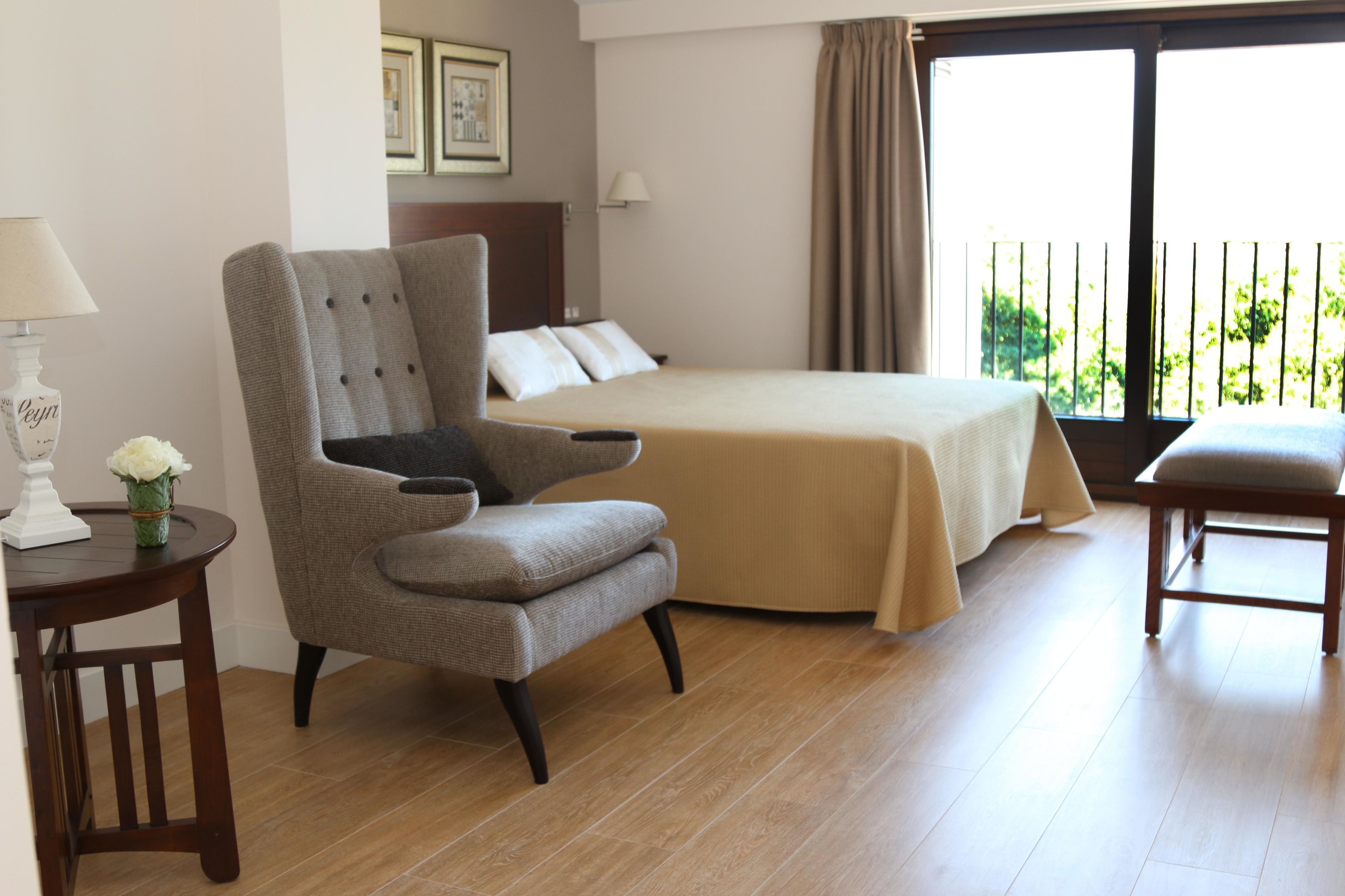 Une chambre d’hôtel avec un grand lit, un coin salon avec un fauteuil, une petite table avec une lampe et une vue sur la verdure par la fenêtre.