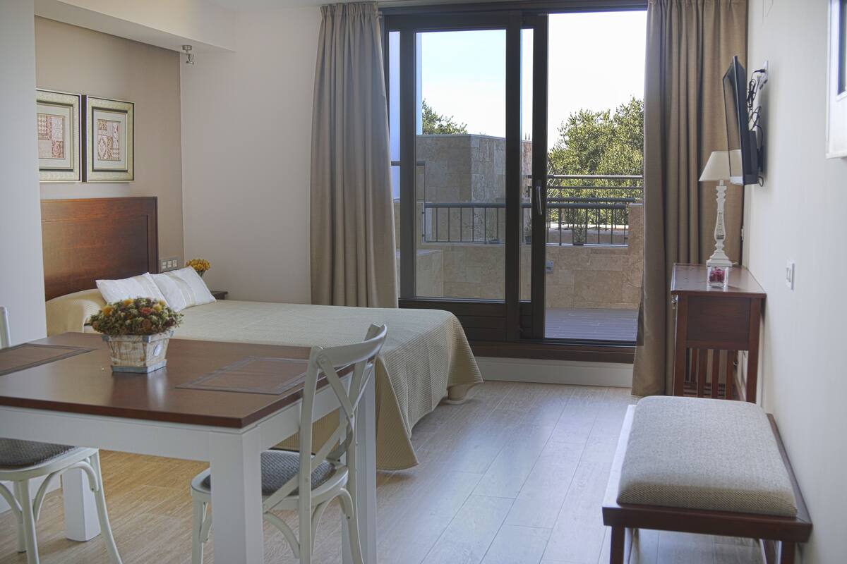 A bedroom with a bed, a desk, a chair, a small table with a vase of flowers, and a bench.