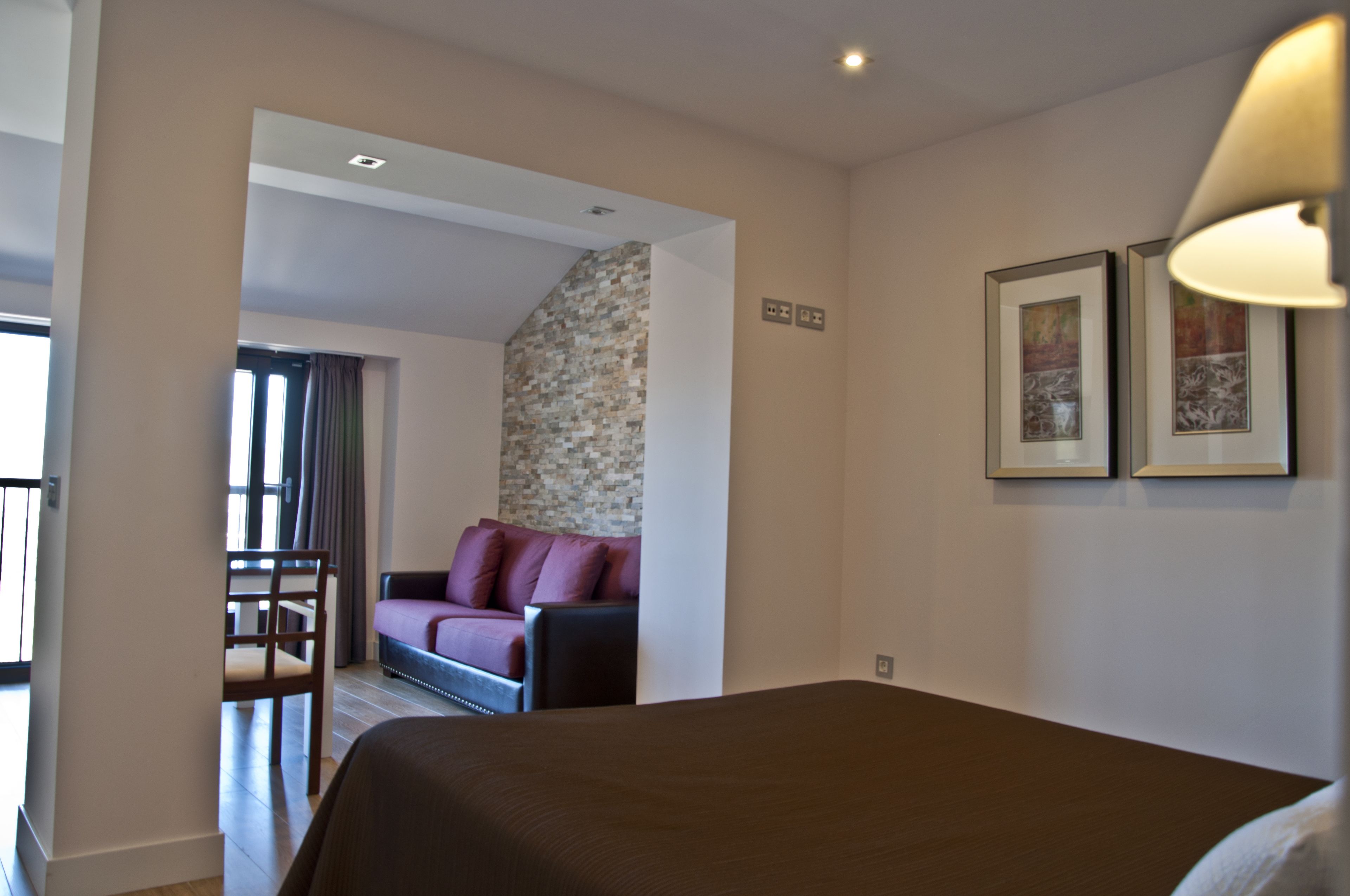 Habitación de hotel con cama, sofá, sillón y una mesita. Hay una ventana con cortinas y una pared con textura.
