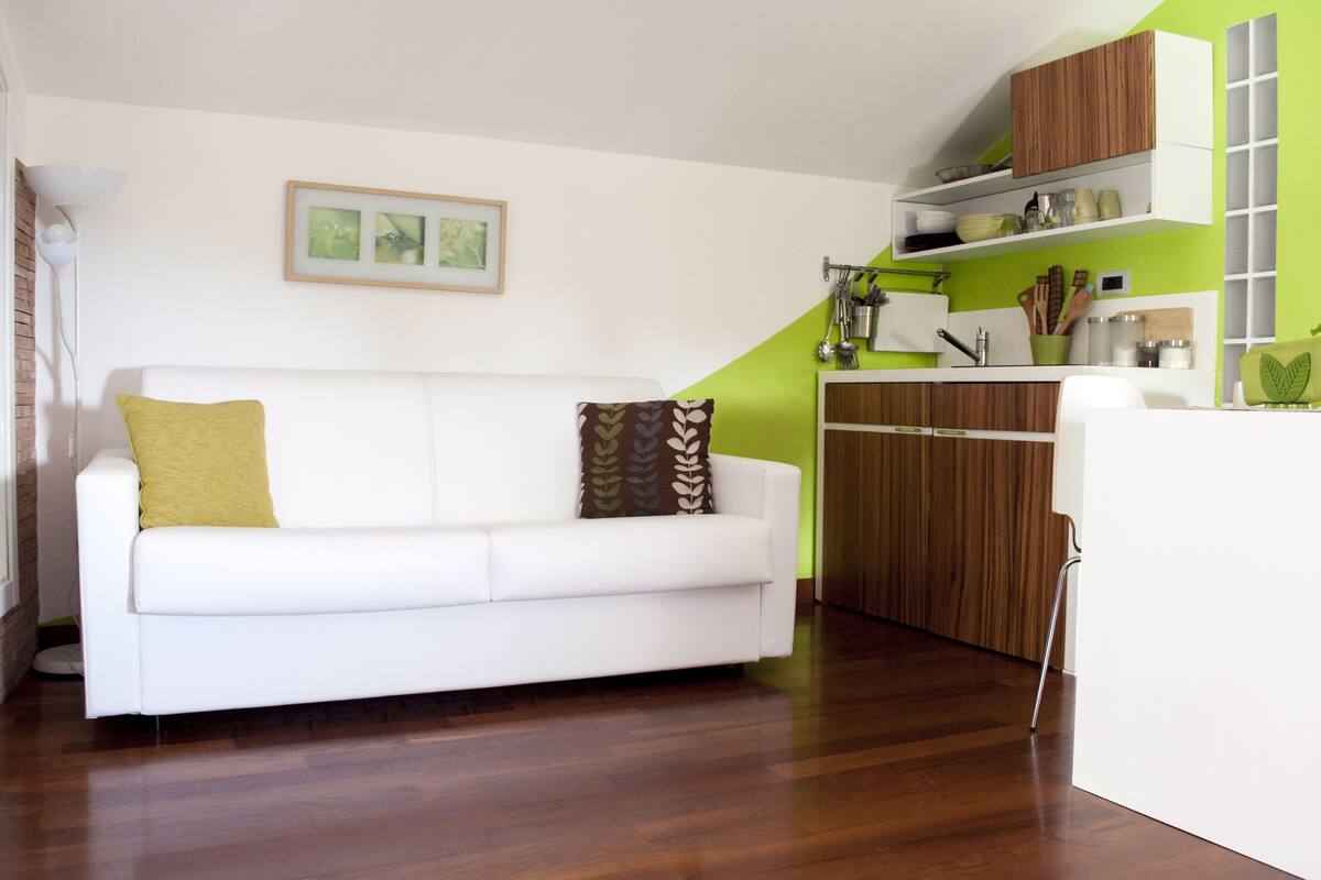 A modern living room with a white sofa, a wooden floor, a green accent wall, and a kitchen area with wooden cabinets and white appliances.