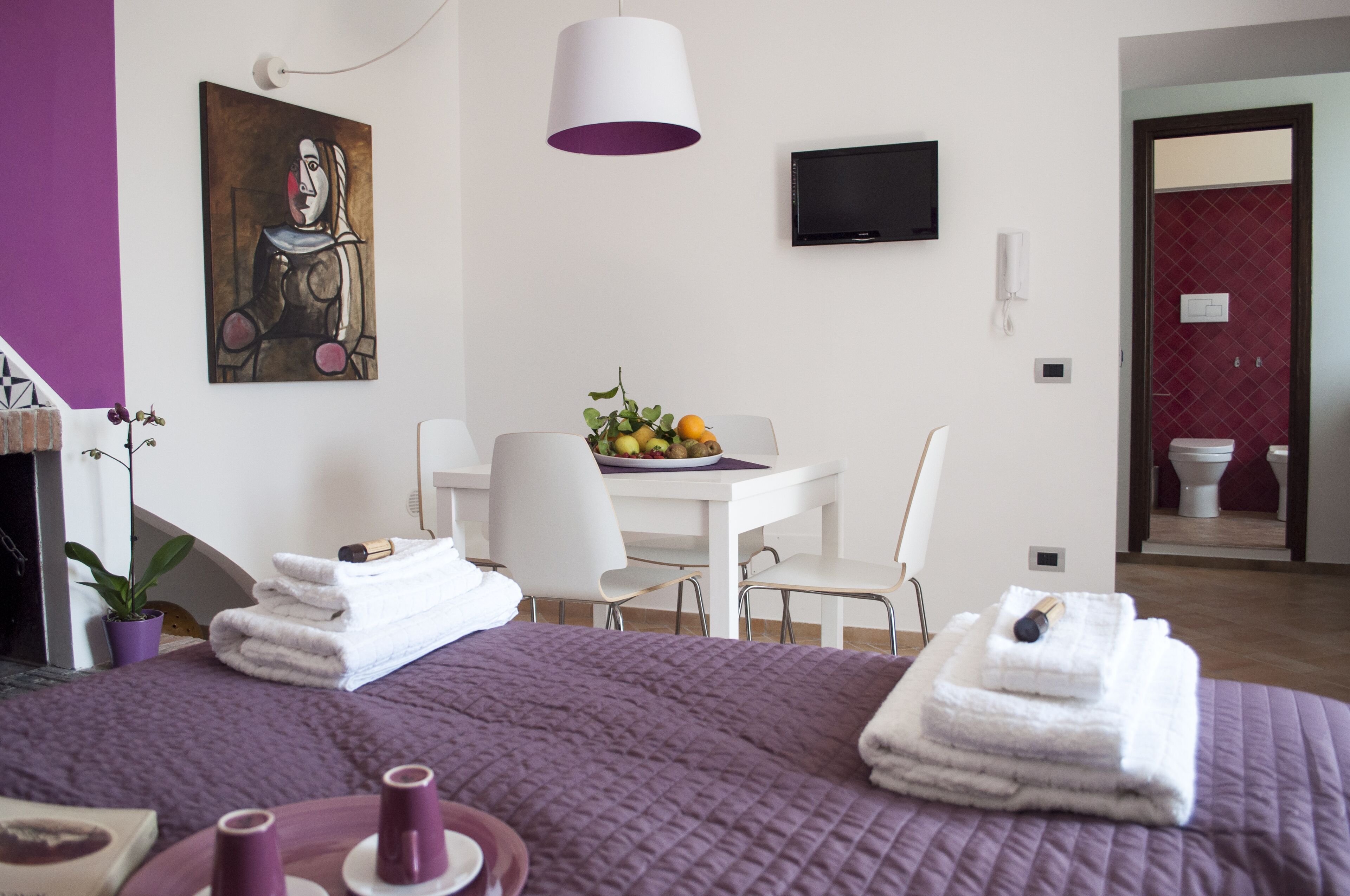 A hotel room with a bed, a desk with a fruit bowl, a TV, and a bathroom visible through an open door.