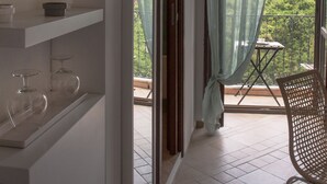 Una habitación con balcón, una silla, estanterías con adornos y vistas al exterior con vegetación.