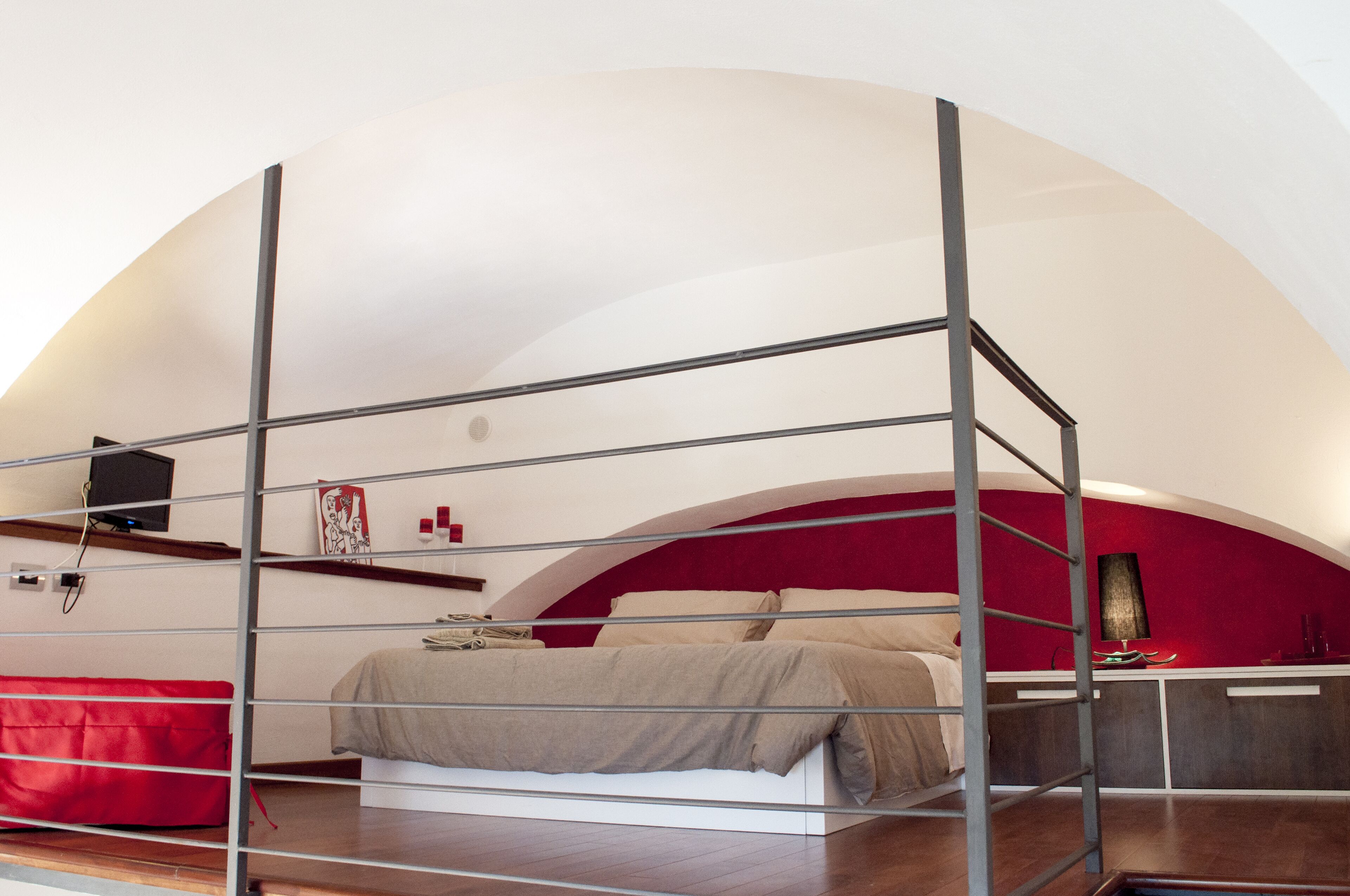 A bedroom with a bunk bed, a red accent wall, a desk with a lamp, and a TV mounted on the wall.