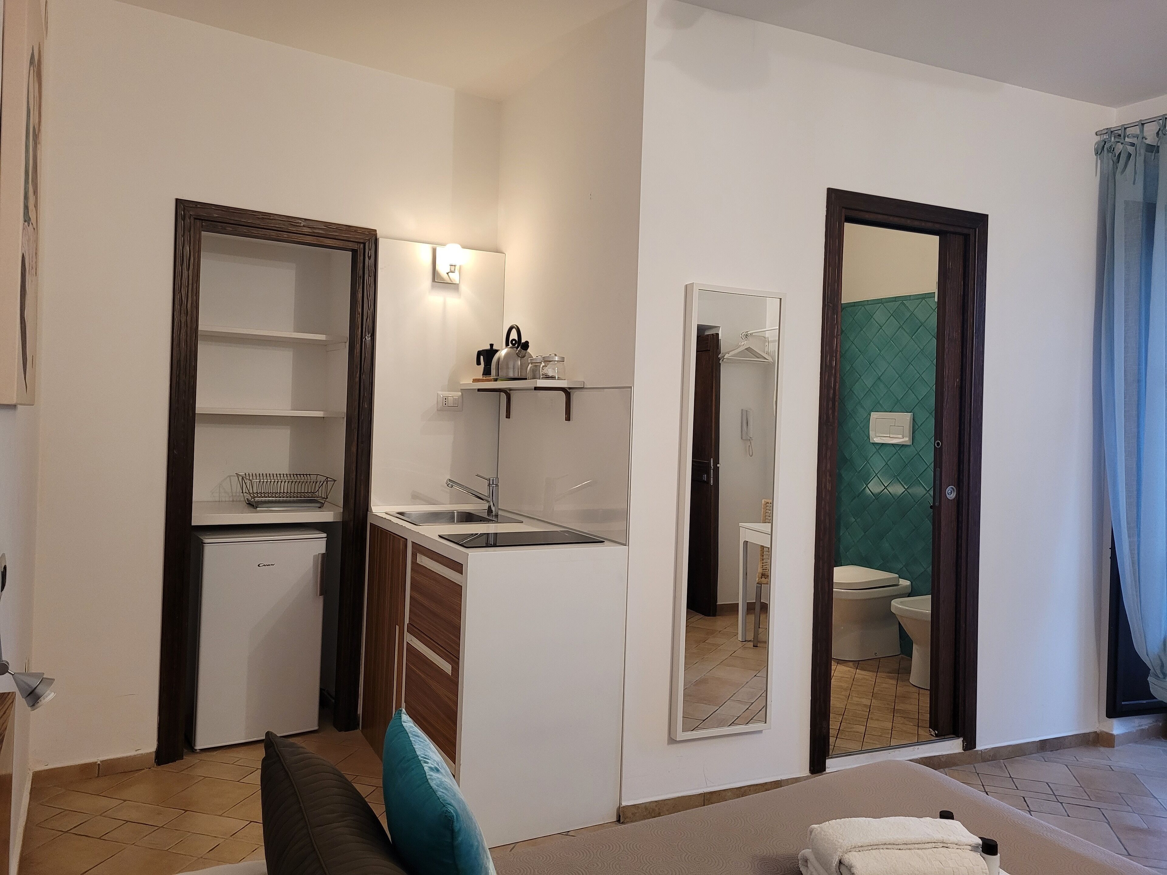 Un espacio habitable compacto con kitchenette, un baño con pared de azulejos verdes y un sofá con un cojín azul aguamarina.