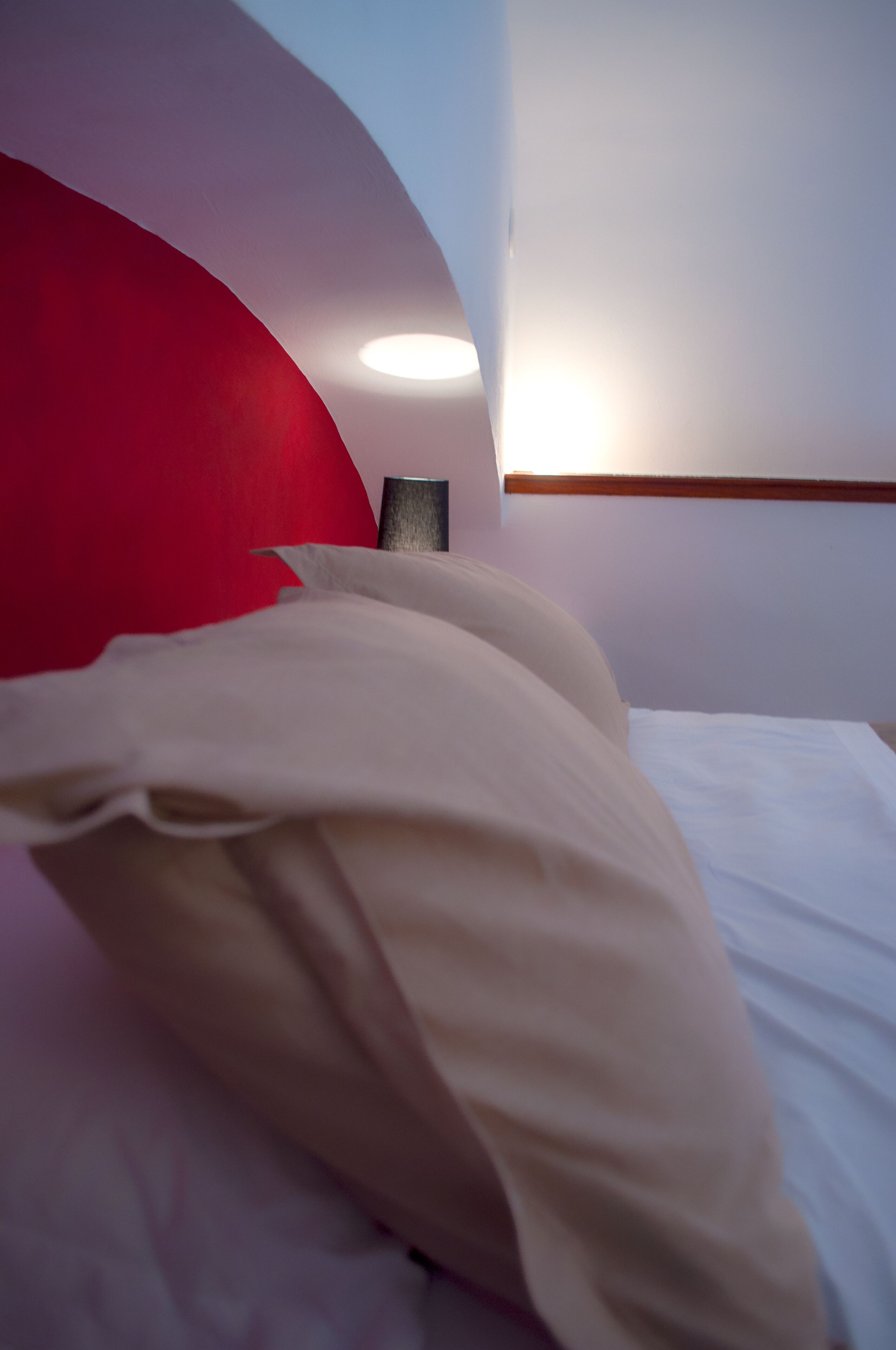 Un lit avec du linge de lit blanc et une tête de lit rouge, une table de chevet et un luminaire fixé au mur.