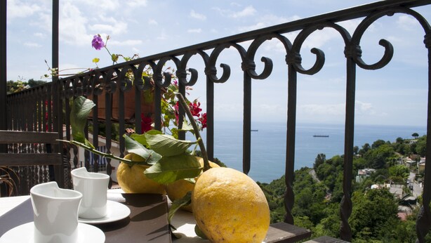 Un balcón con una mesa dispuesta para dos, una fruta amarilla grande y vista al mar.