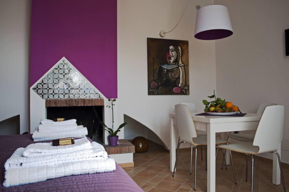 A modern hotel room with a purple accent wall, a fireplace, a dining area with white chairs, and a table with fruit.