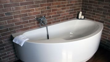 Une baignoire blanche, autoportante, avec une serviette, une pomme de douche et des bougies, le tout placé devant un mur de briques.