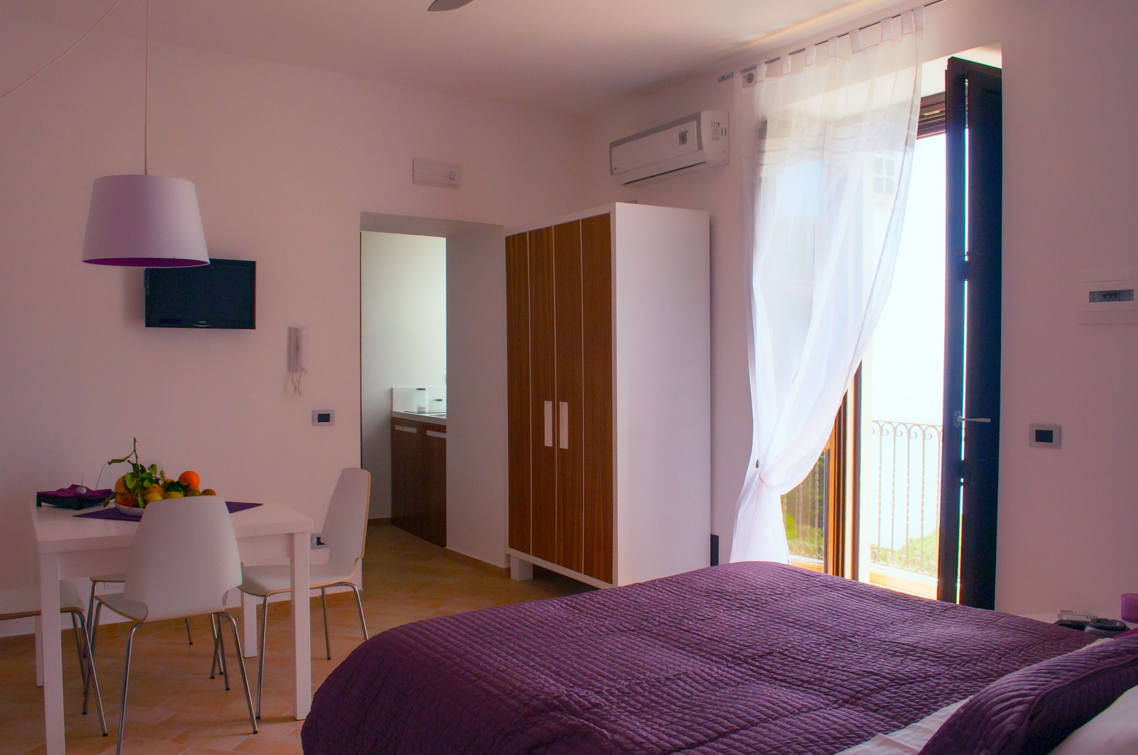 A bedroom with a bed, a desk with chairs, a wardrobe, and a balcony.