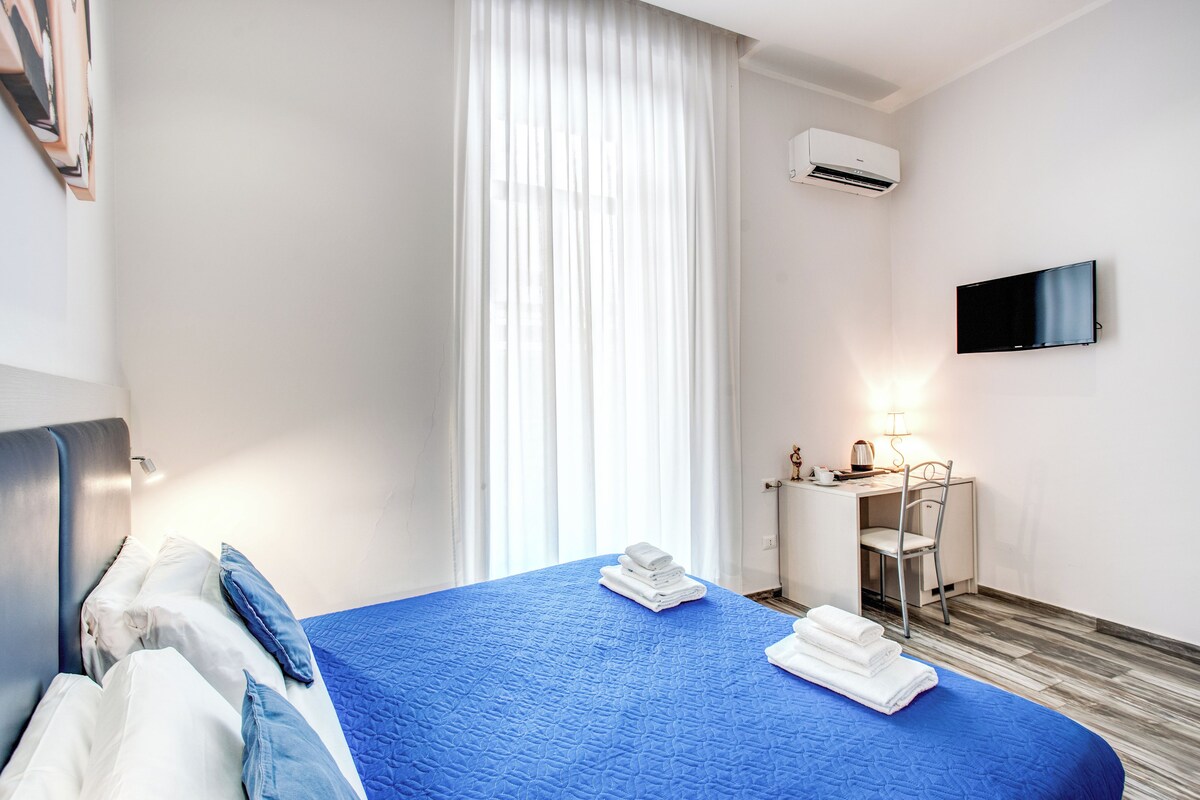A hotel room with a bed, a desk, a chair, a TV, and an air conditioning unit.