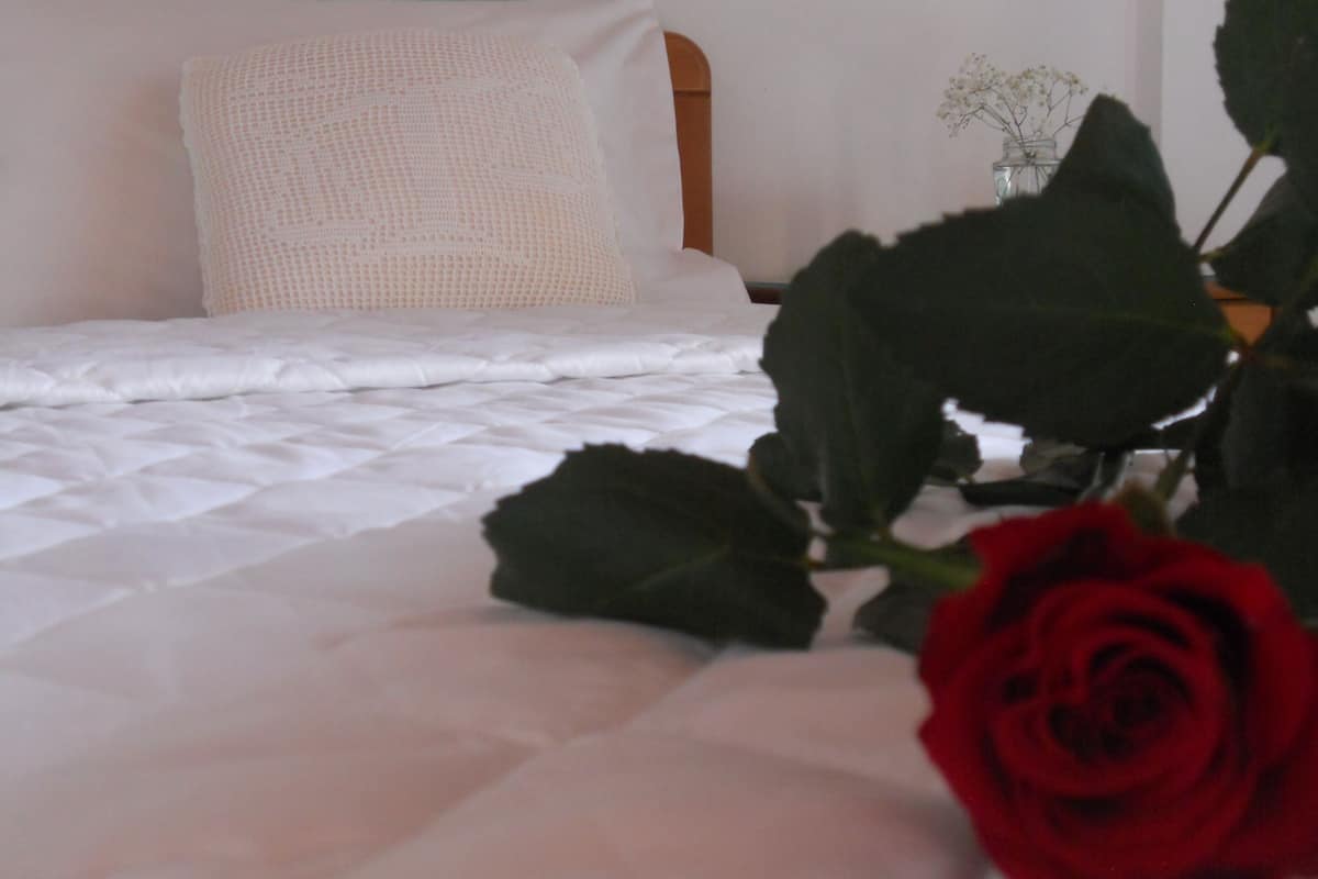 Uma cama com cobertura branca acolchoada, um travesseiro branco e uma rosa vermelha em primeiro plano.