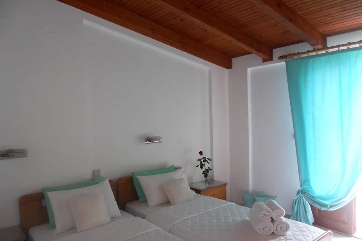 Quarto de hotel com duas camas, vigas de madeira no teto, uma pequena mesa de cabeceira com um vaso e uma janela com cortina.