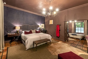 A bedroom with a large bed, a wooden bench, a chandelier, a red robe, and a vintage bathtub.