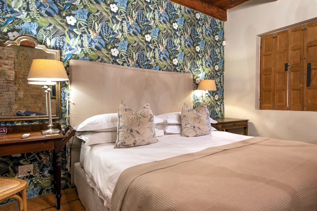 A bedroom with a floral wallpaper, a wooden headboard, a bed with white linens, two bedside lamps, and a wooden wardrobe.