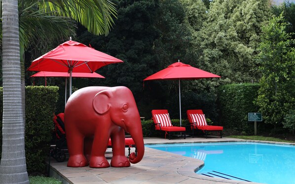 Un área de piscina con sombrillas rojas, una estatua grande de elefante y sillones reclinables rojos.