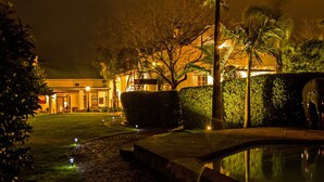 A nighttime view of a hotel courtyard with a swimming pool, well-maintained hedges, and a large building with illuminated windows. - Avondrood Guest House (Franschhoek)
