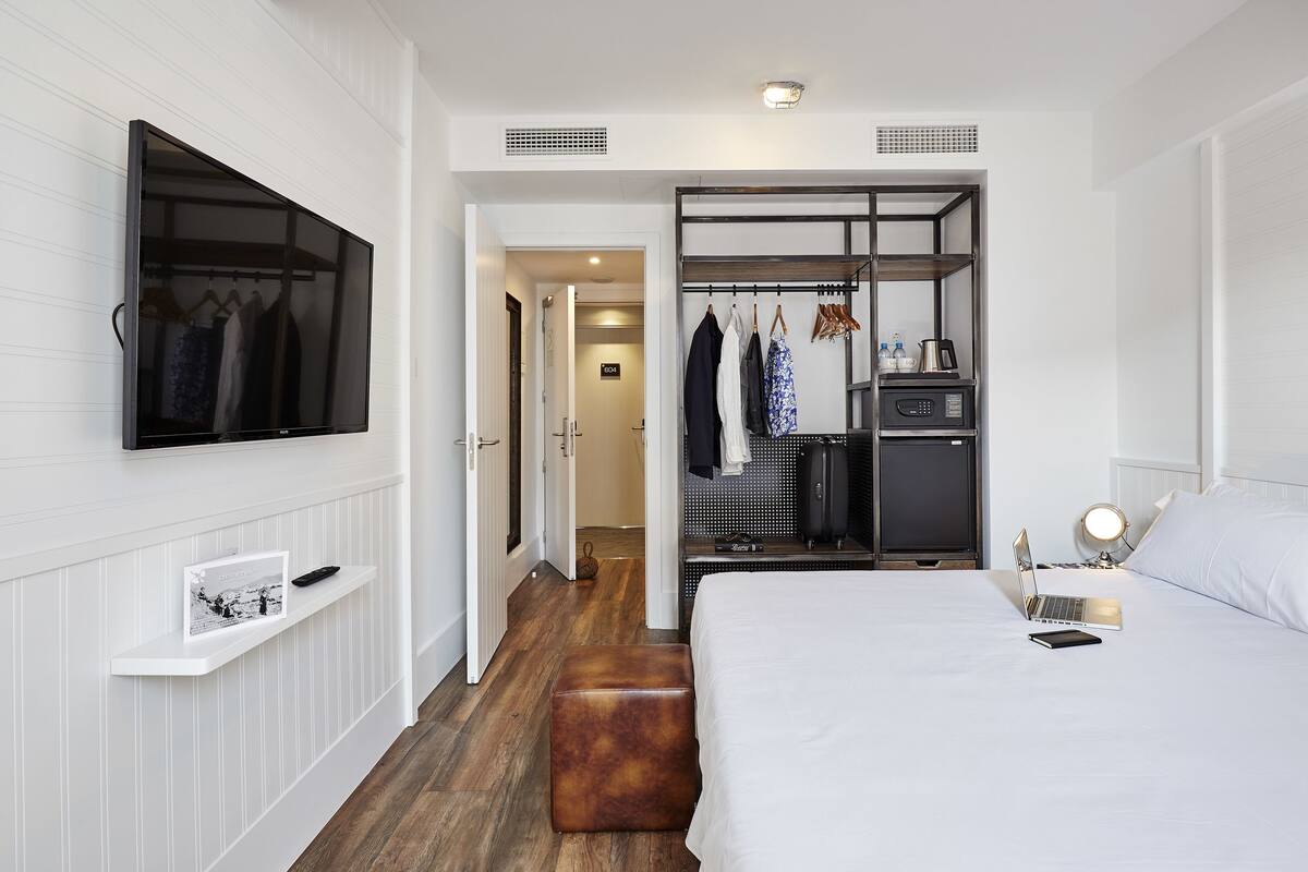 Une chambre d’hôtel moderne avec un grand lit, un téléviseur fixé au mur, une armoire avec des vêtements suspendus et un petit bureau avec une lampe.