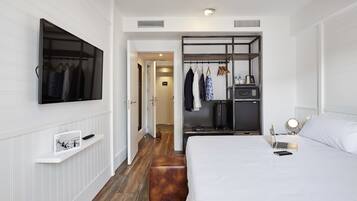 A modern hotel room with a large bed, a TV mounted on the wall, a wardrobe with hanging clothes, and a small desk with a lamp.