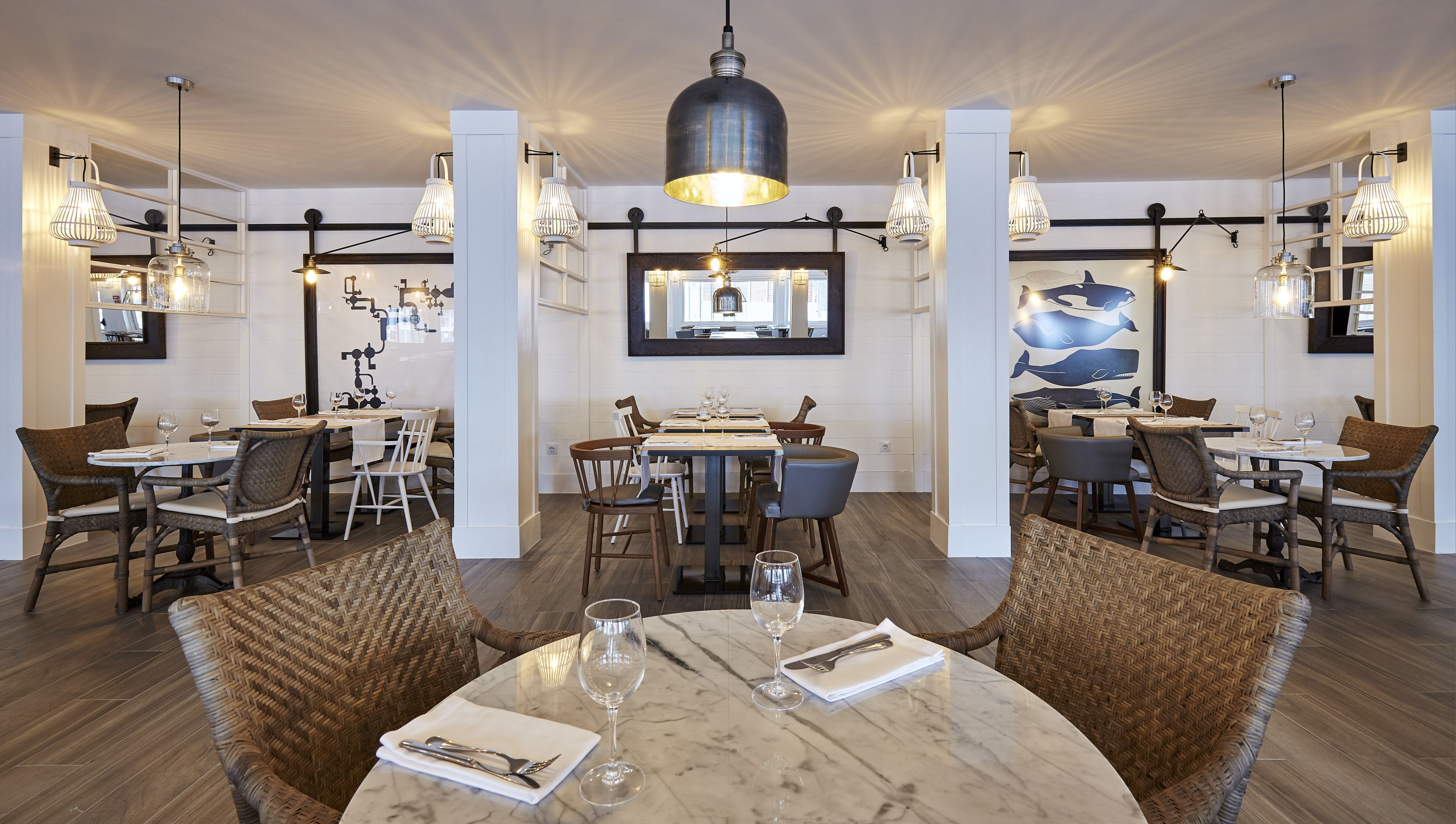 Een restaurantinterieur met ronde marmeren tafels, rieten stoelen en hanglampen.