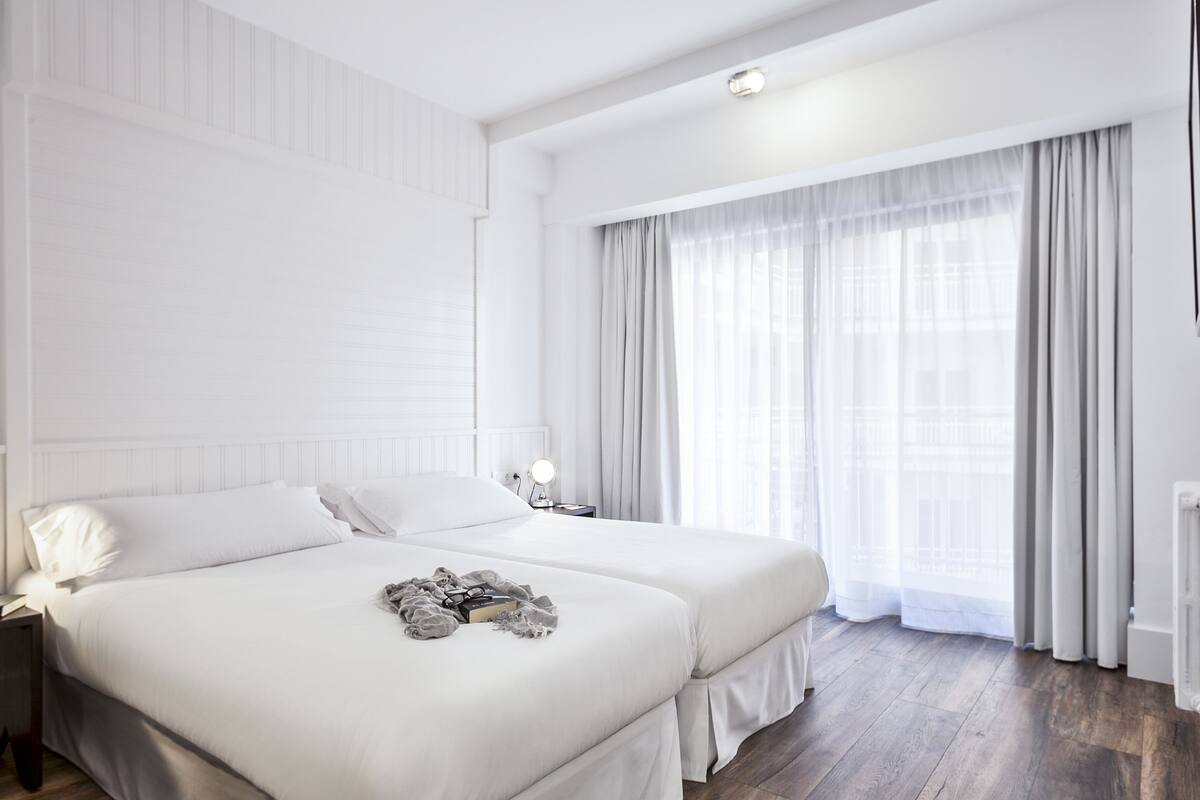 Une chambre d’hôtel moderne avec deux lits, une grande fenêtre avec des rideaux légers, une table de chevet avec une lampe et un radiateur.