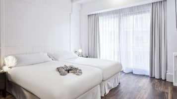 Habitación de hotel moderna con dos camas, un ventanal grande con cortinas translúcidas, una mesita de noche con lámpara y un radiador.